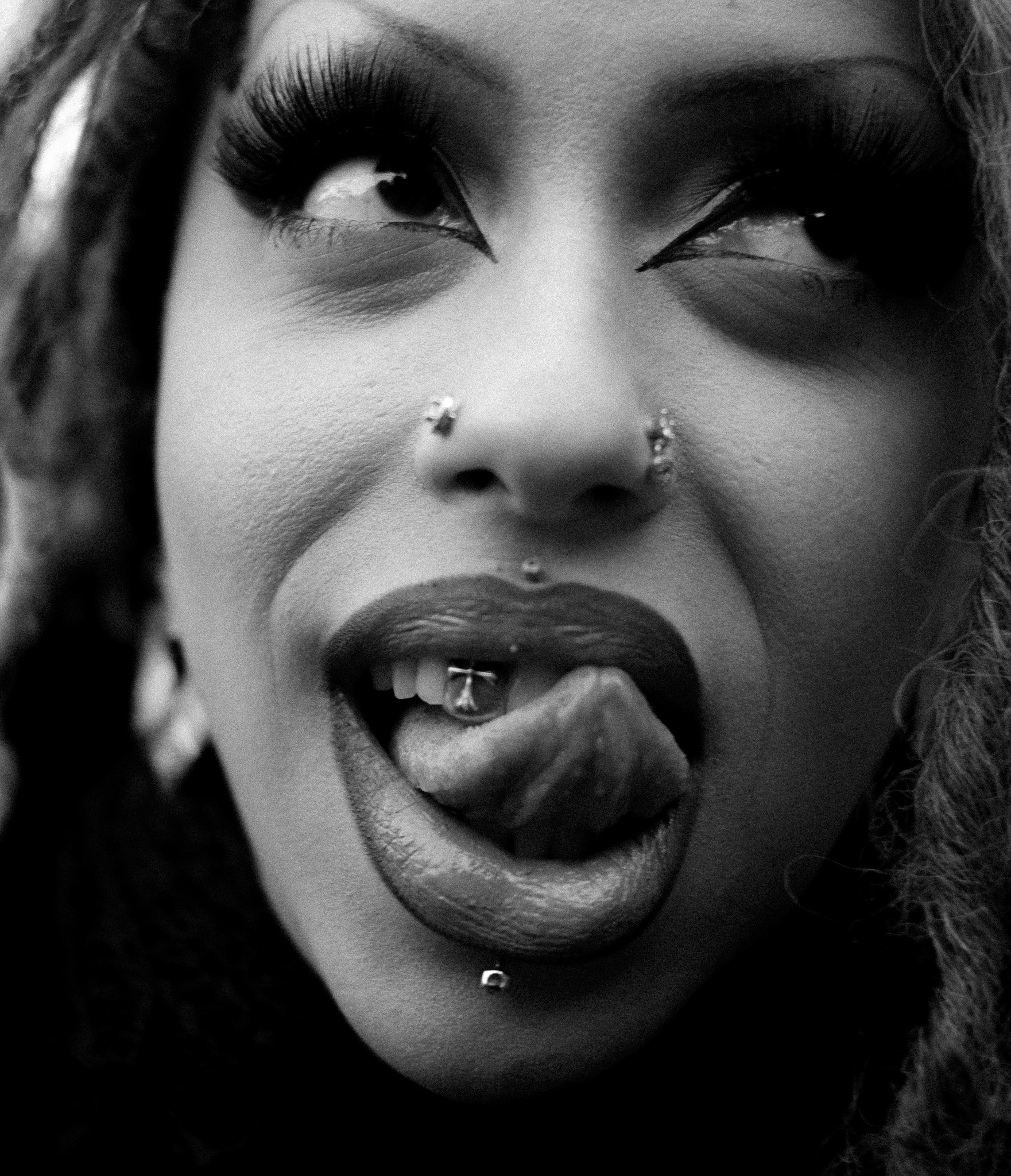 Close-up black and white photo of a woman's face with makeup, nose piercings, lips slightly parted, and curly hair partially visible.