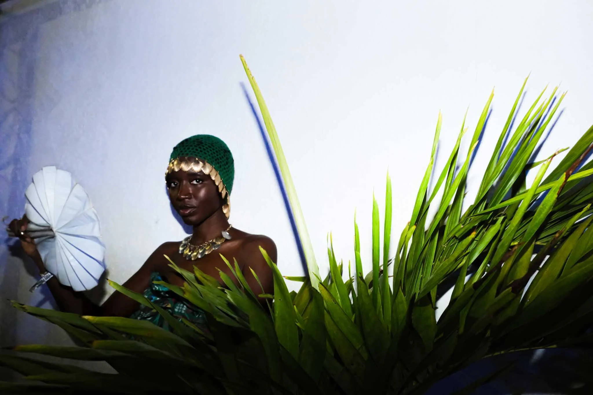 A woman with dark skin wearing traditional attire with a green headpiece and jewelry, holding a white folded paper fan, standing behind large green leaves or grass, against a plain white wall.