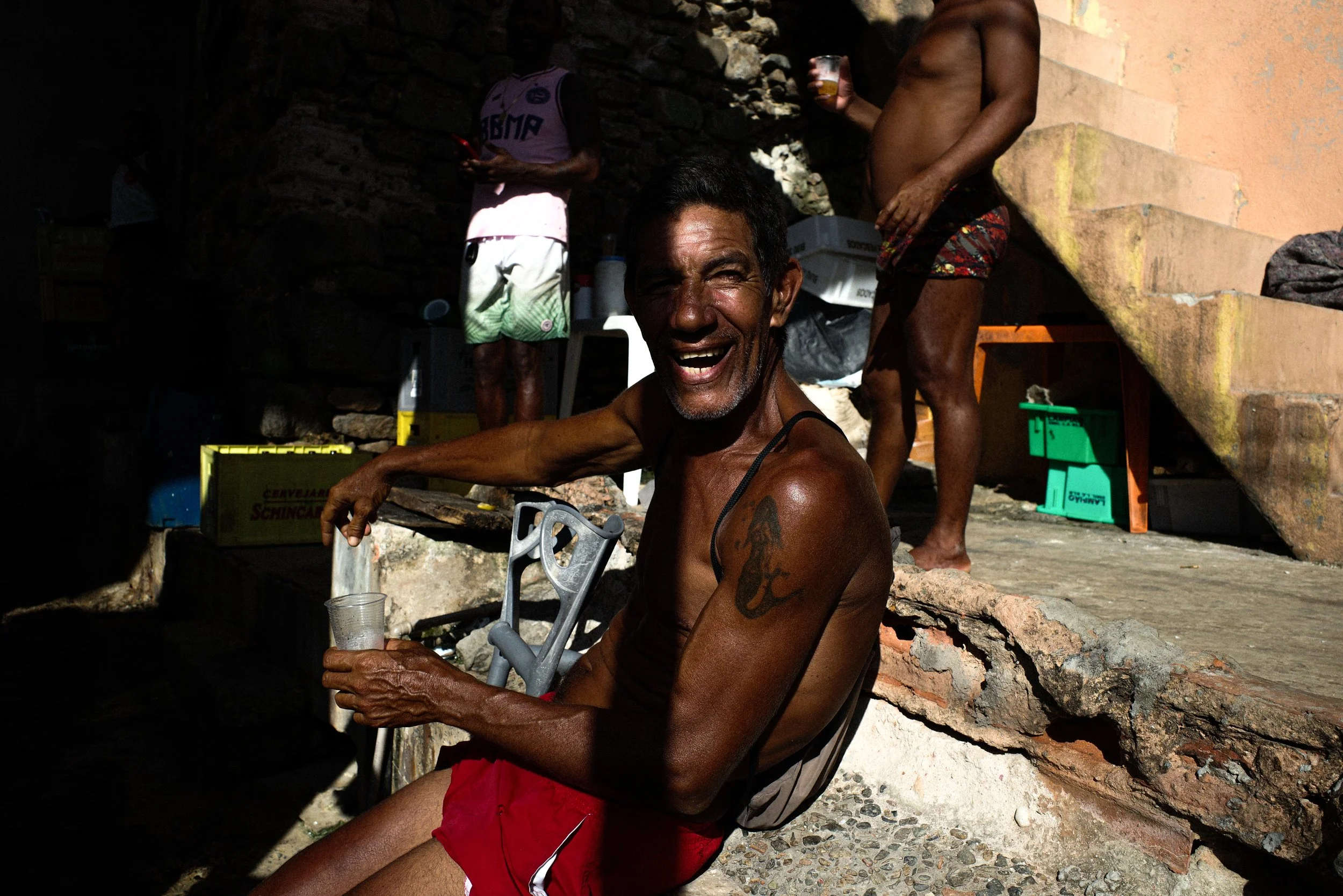 A smiling man with dark skin and tattoos on his arm, sitting outdoors in a sunny area, holding a glass of drink. In the background, two other people are near a rock wall, one of them shirtless, the other wearing a tank top and holding a drink, with various boxes and containers around.
