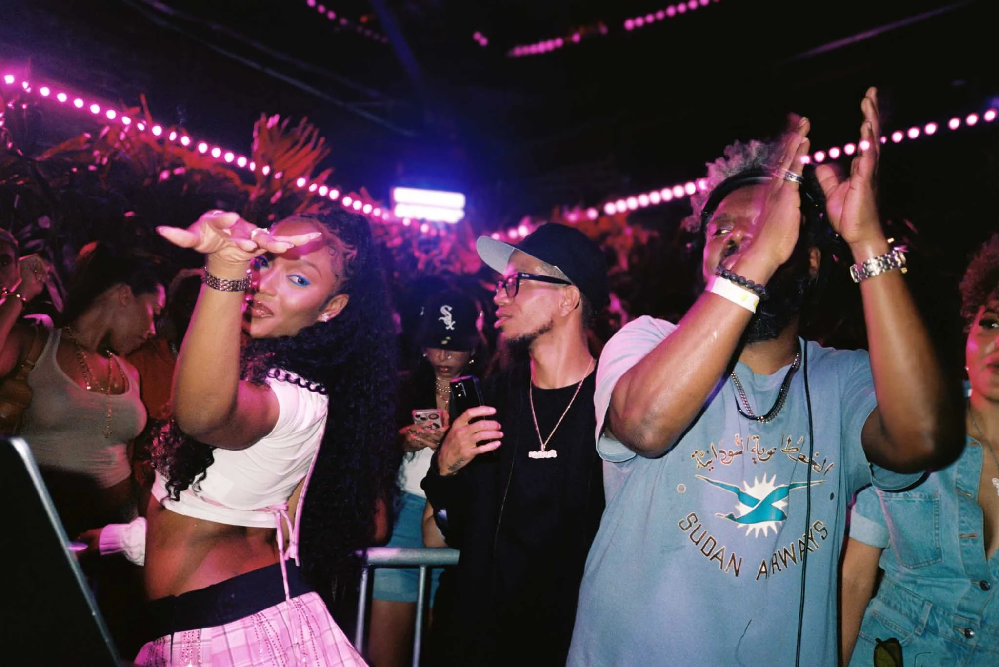 A group of people at a nightclub dancing and enjoying the music under pink neon lights. Women are wearing stylish clothes and jewelry, and men are casually dressed with accessories. The atmosphere appears lively and festive.