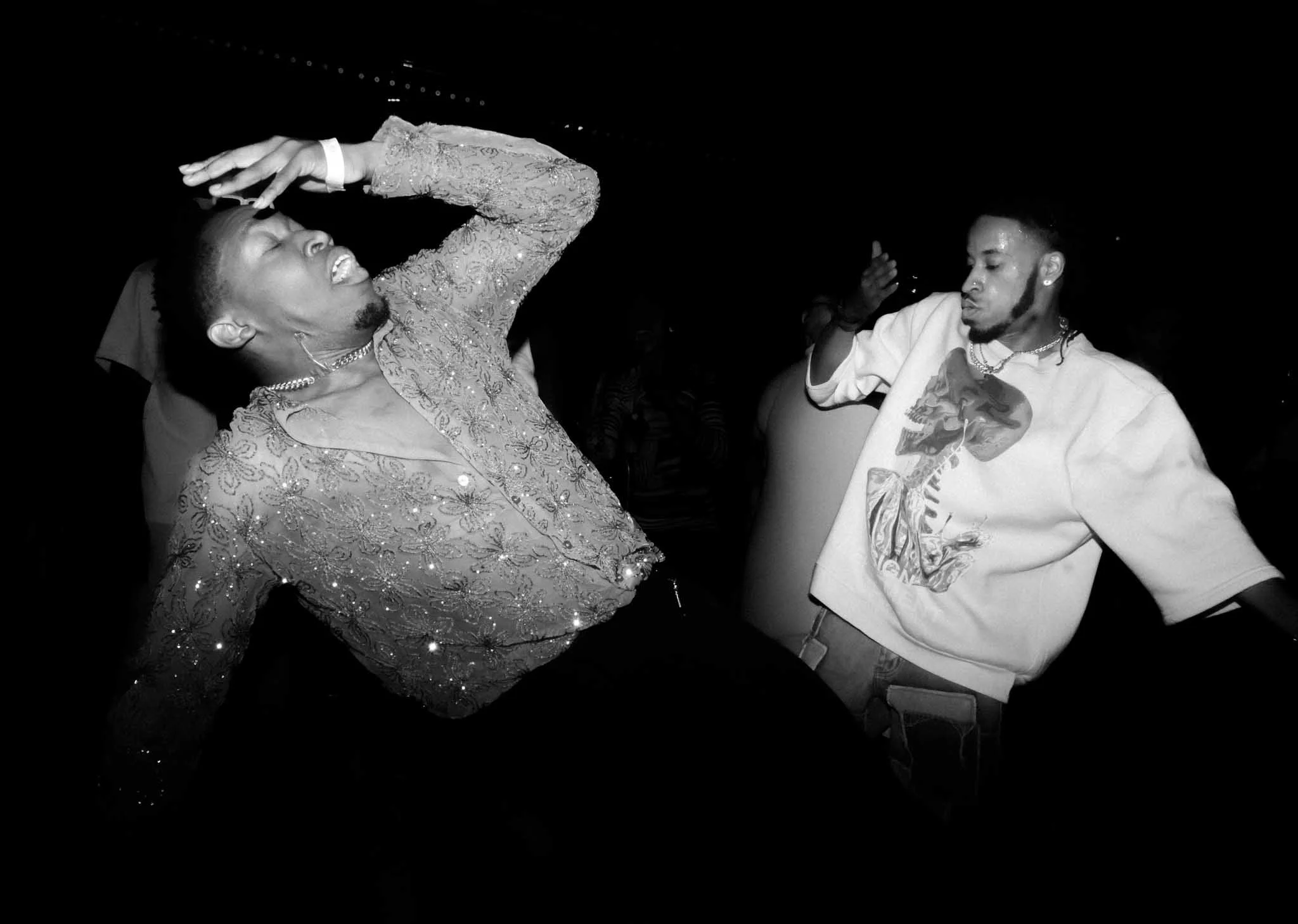 Two individuals dancing at a party or club, with one person wearing a shiny, patterned shirt and the other wearing a white graphic T-shirt, both appearing to enjoy the moment.