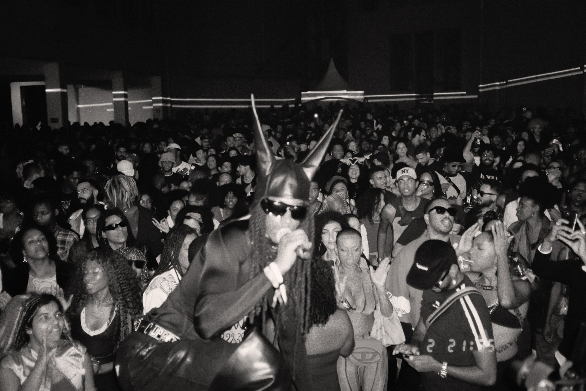 A large crowd of people at an indoor event, with a person in the foreground wearing a jester hat and sunglasses, holding a phone, and others taking photos or videos in a lively, crowded atmosphere.