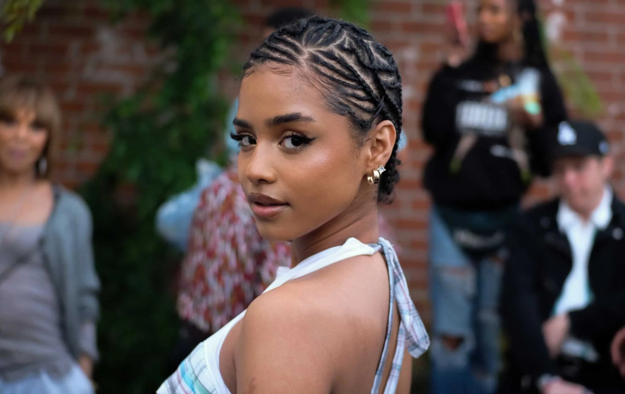 Close-up of a woman with braids, looking over her shoulder at the camera, with friends blurred in the background.