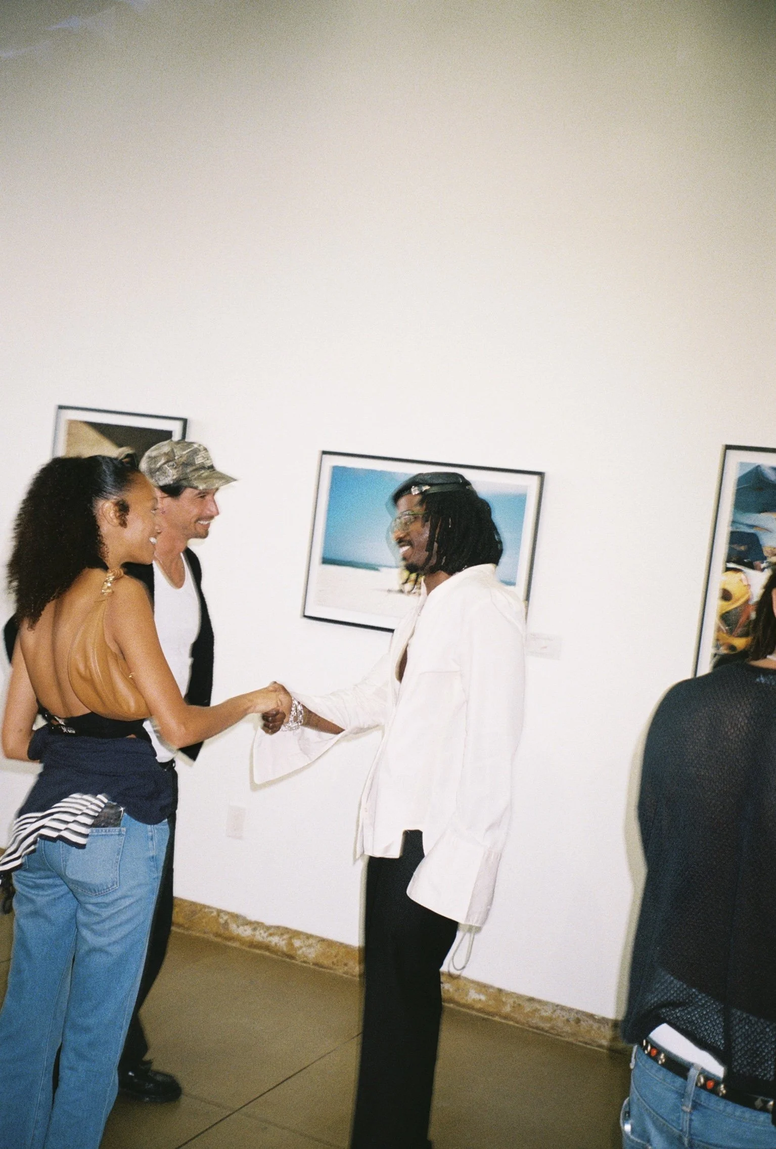 A man and a woman are shaking hands and smiling while another man stands nearby, in an art gallery with framed photographs on the wall.