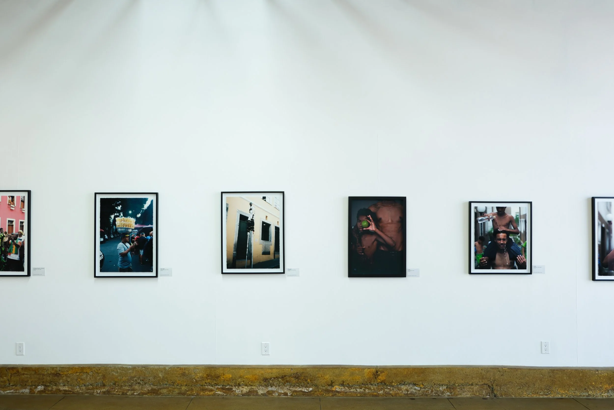 A series of framed photographs displayed on a white gallery wall, showing diverse scenes including people in urban environments, individuals holding objects, and street life, with small descriptive labels beneath each frame.