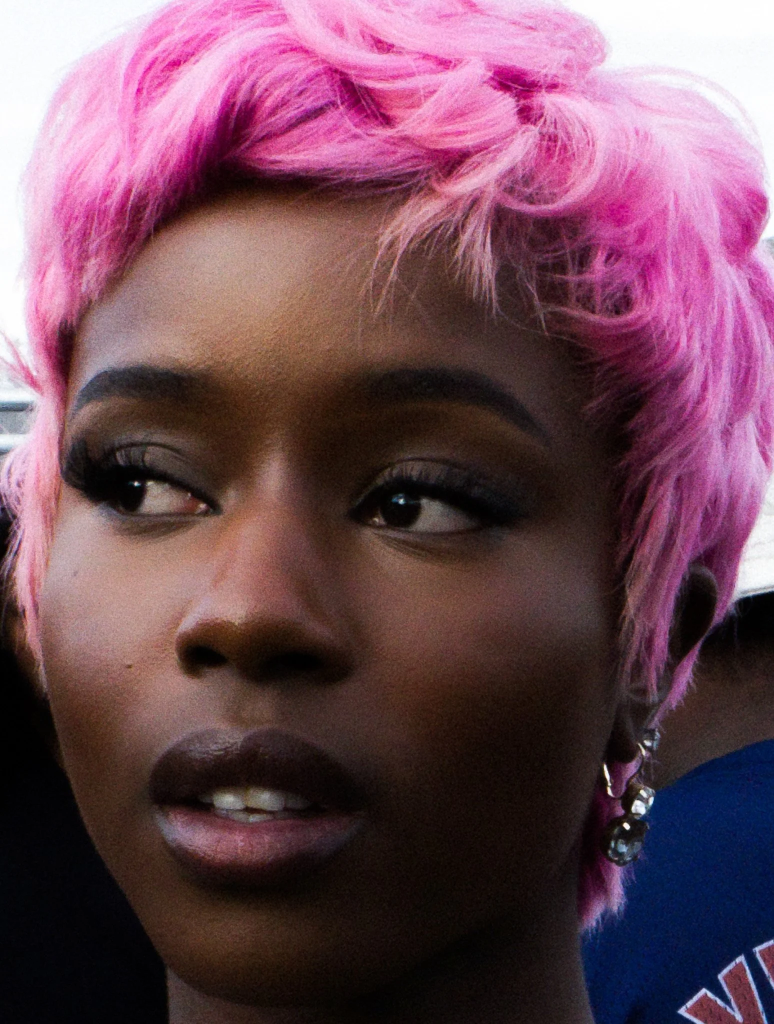 Close-up of a woman with pink styled hair, dark eyebrows, and makeup, celebrating with her mouth open and showing her teeth.