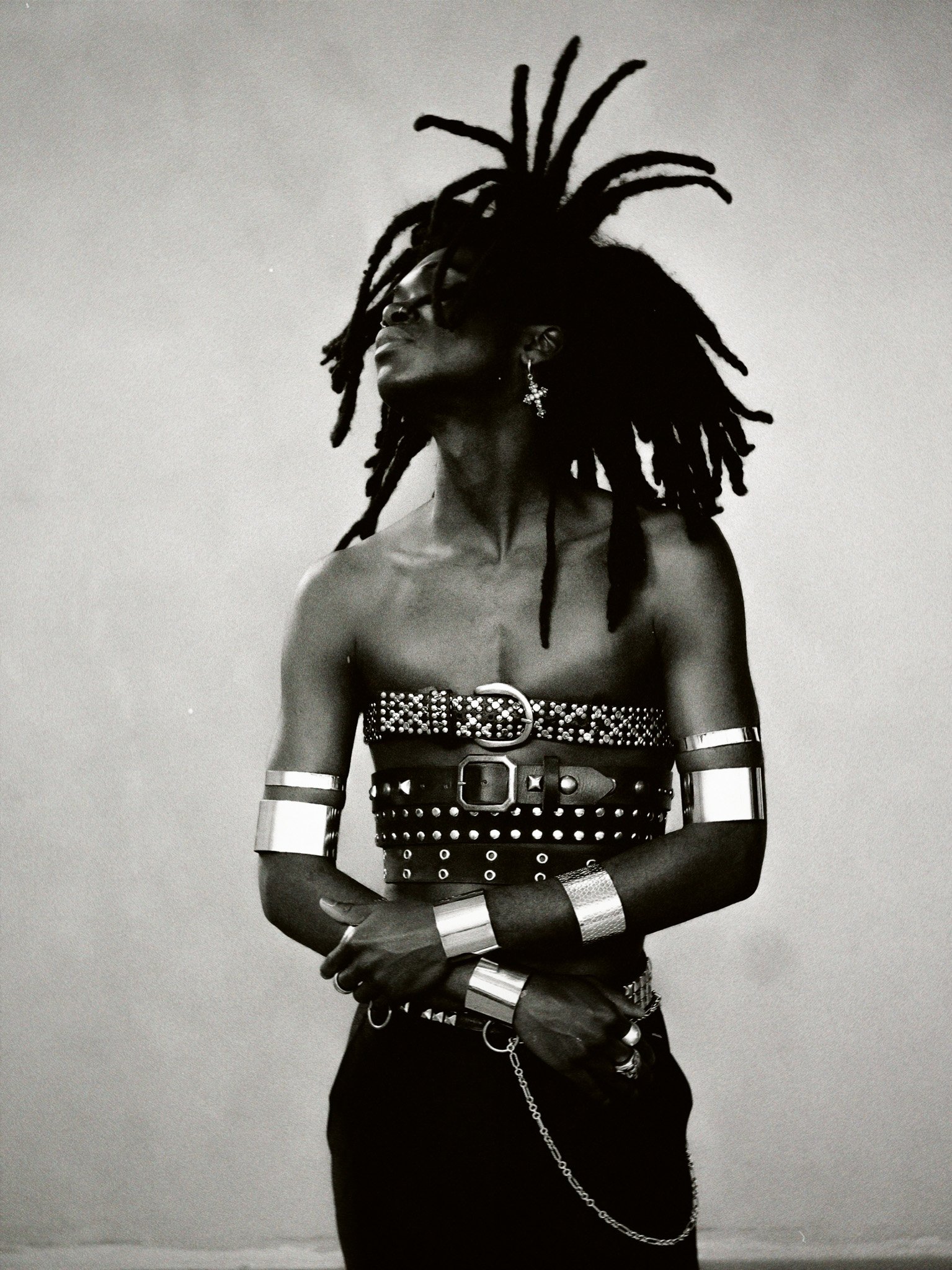 A black and white photo of a woman with dreadlocks, wearing multiple metallic arm cuffs, a studded belt, dangling earrings, and rings, posing against a plain light background.