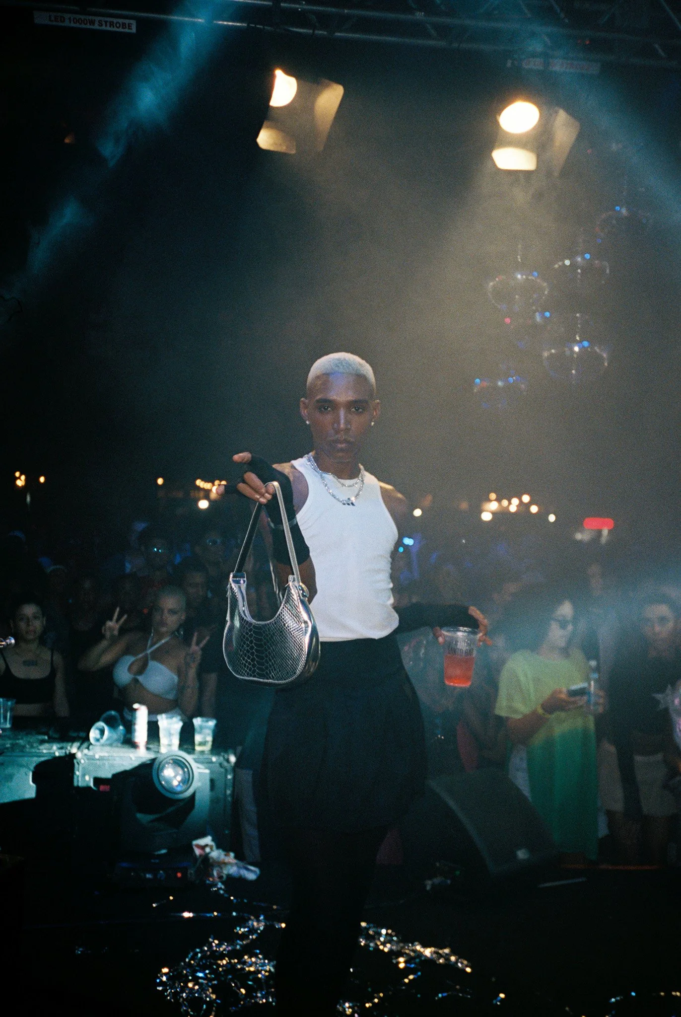 A woman with short blonde hair wearing a white tank top and black pants is holding a silver handbag and a plastic cup with a red beverage in a nightclub with disco balls and bright lights. In the background, several people are dancing and taking pict