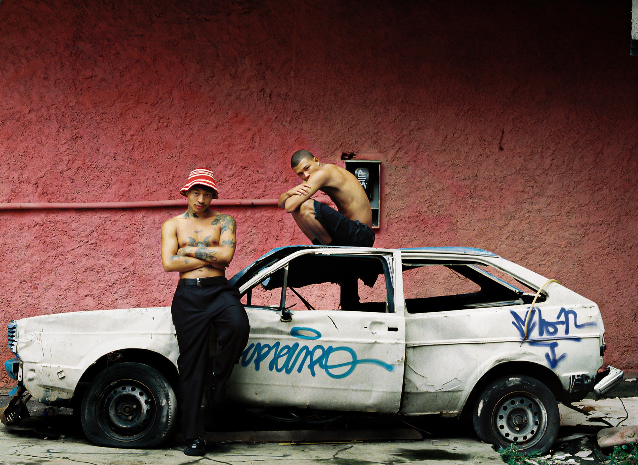 Two shirtless young men with tattoos posing by an old, graffiti-covered white car against a red textured wall. One wears a red striped bucket hat, the other crouches on the car roof.