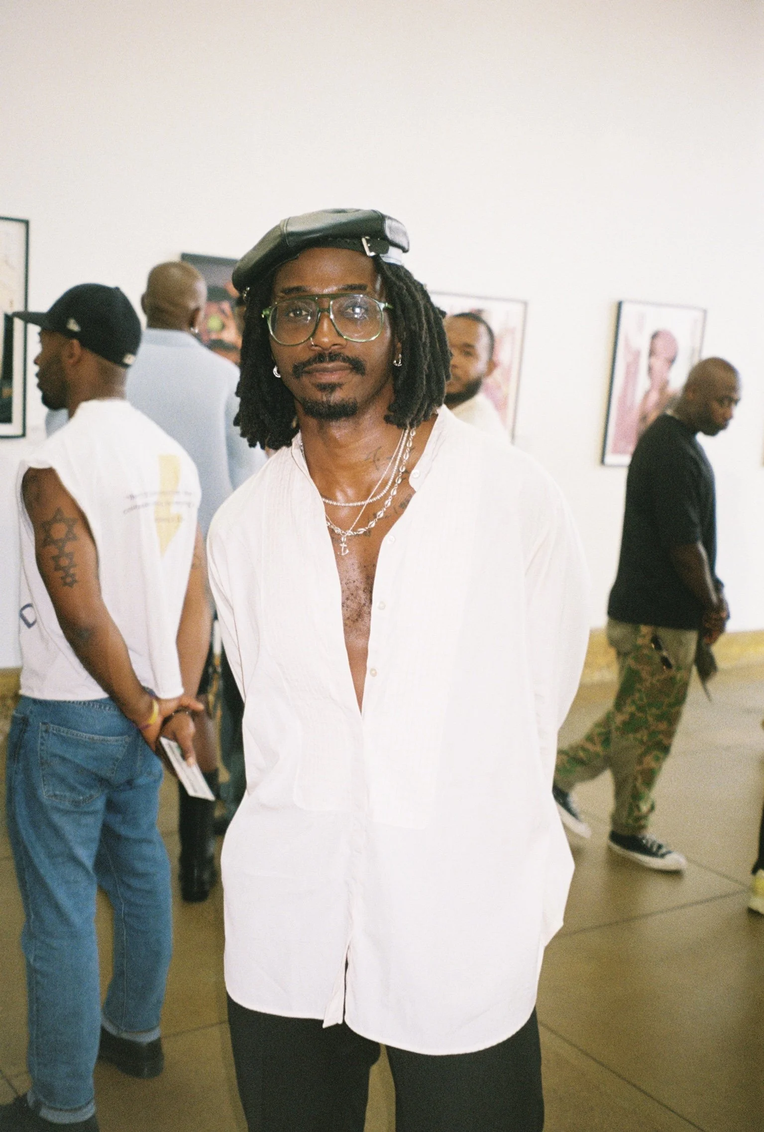 A man with glasses, earrings, necklace, and dreadlocks standing in an art gallery, with people in the background looking at artwork.