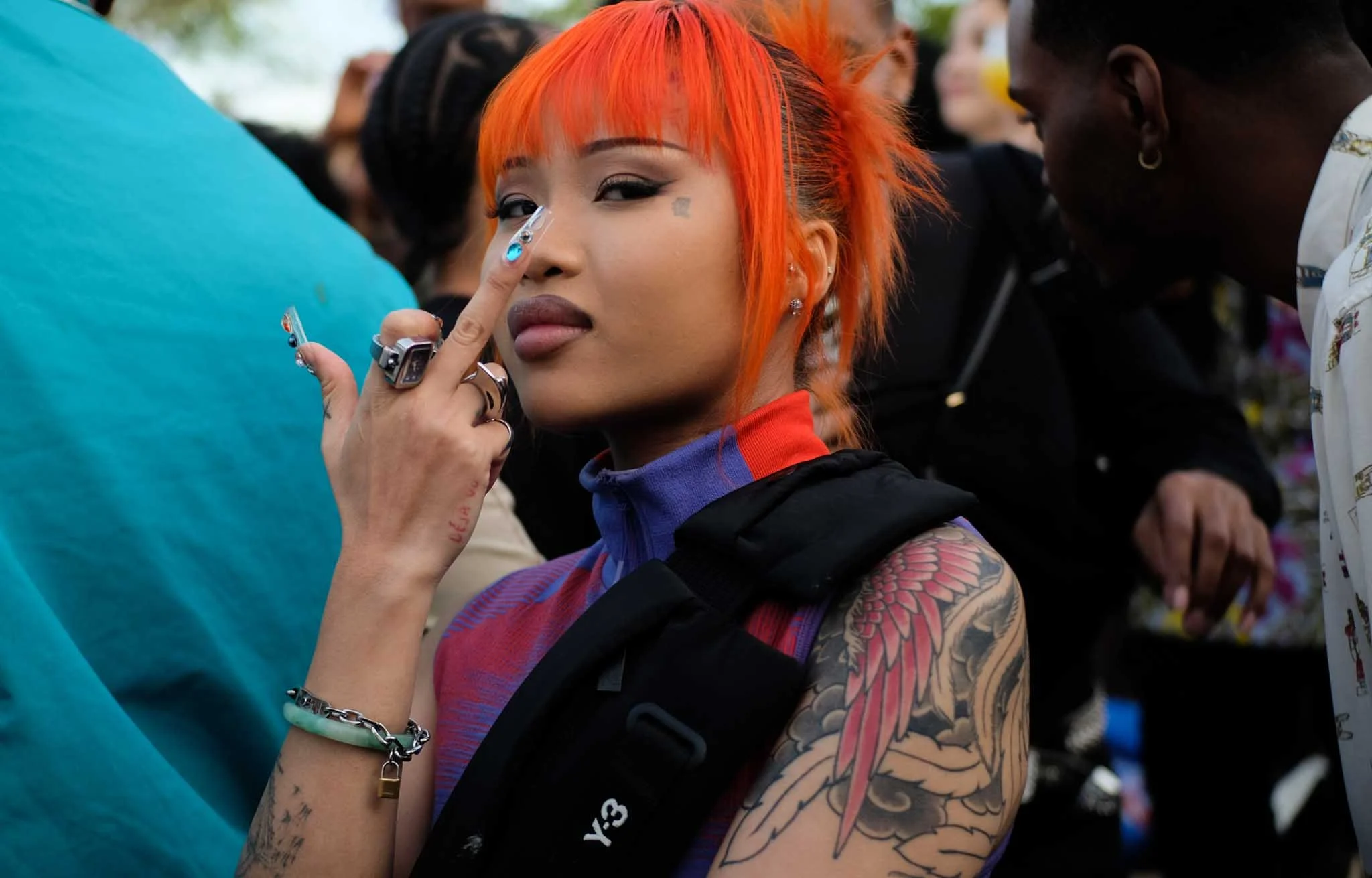 A woman with bright orange hair and tattoos on her arm holds a finger near her eye while looking at the camera, surrounded by people in a crowded outdoor setting.
