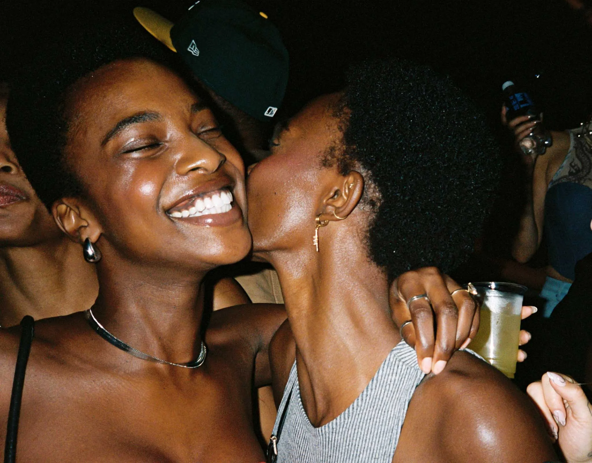 Two women sharing a close, joyful moment, smiling, with heads touching, in a dark setting, wearing casual clothing and accessories.
