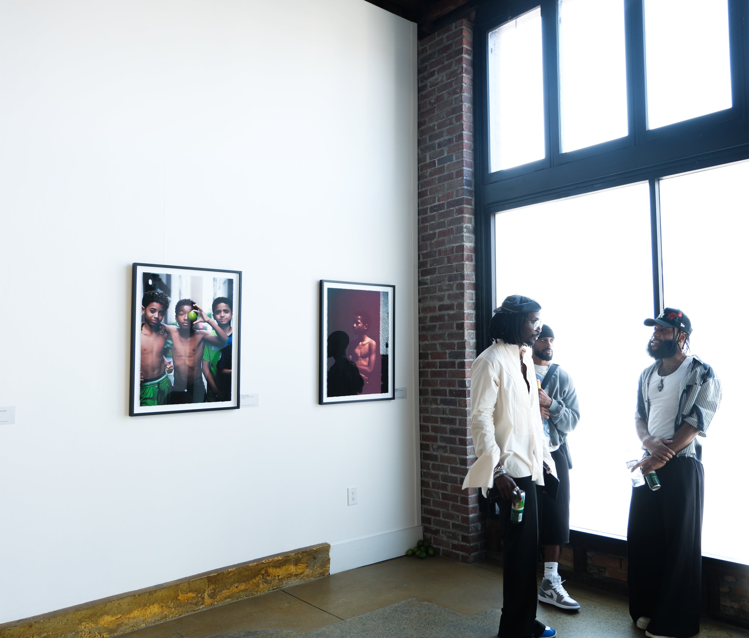 People standing near a white wall with framed photos, inside an art gallery or exhibition space with a large window and exposed brick wall.