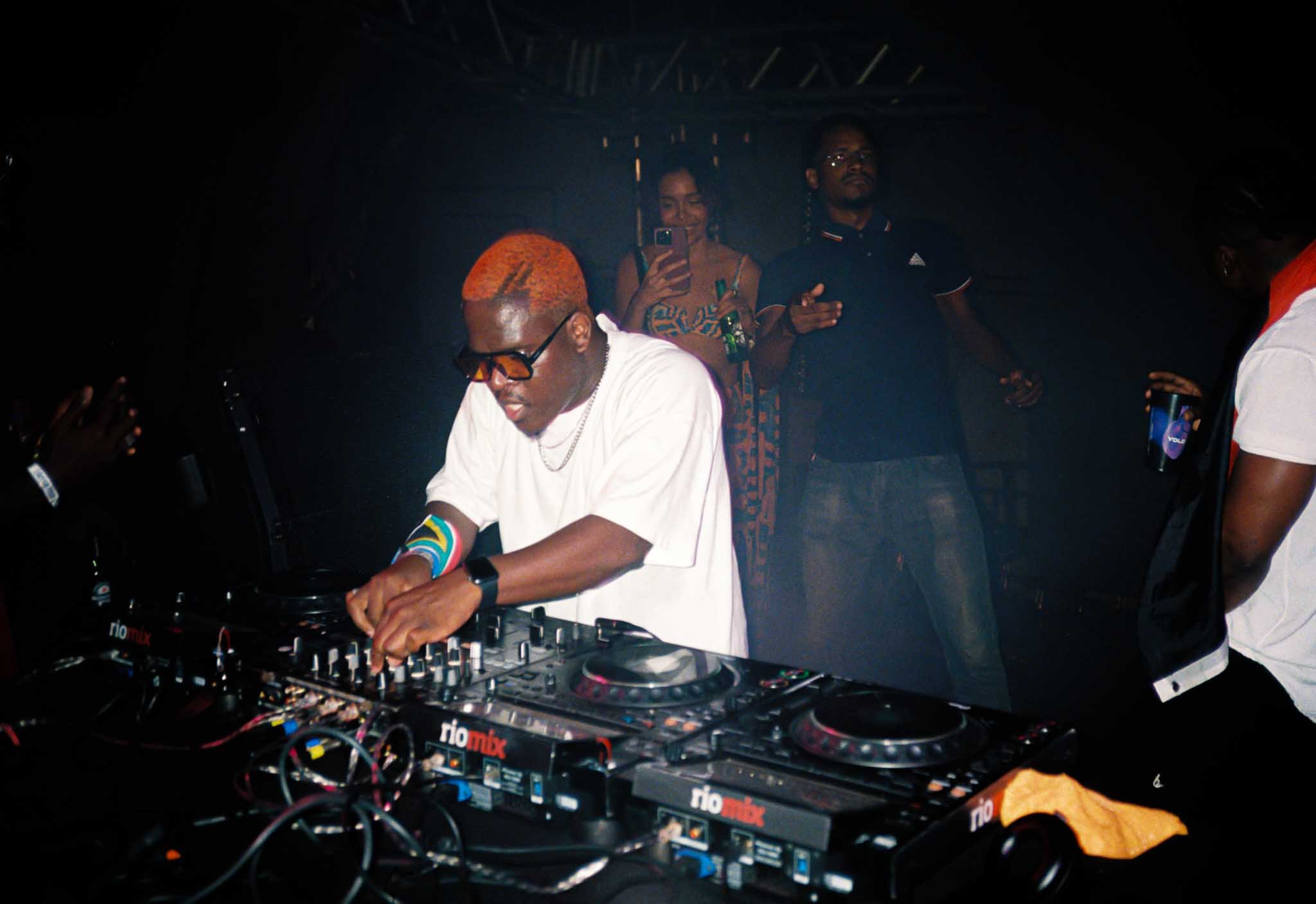 A DJ with orange hair and sunglasses is performing at a party with a DJ mixer and equipment, surrounded by people enjoying the event in a dimly lit space.