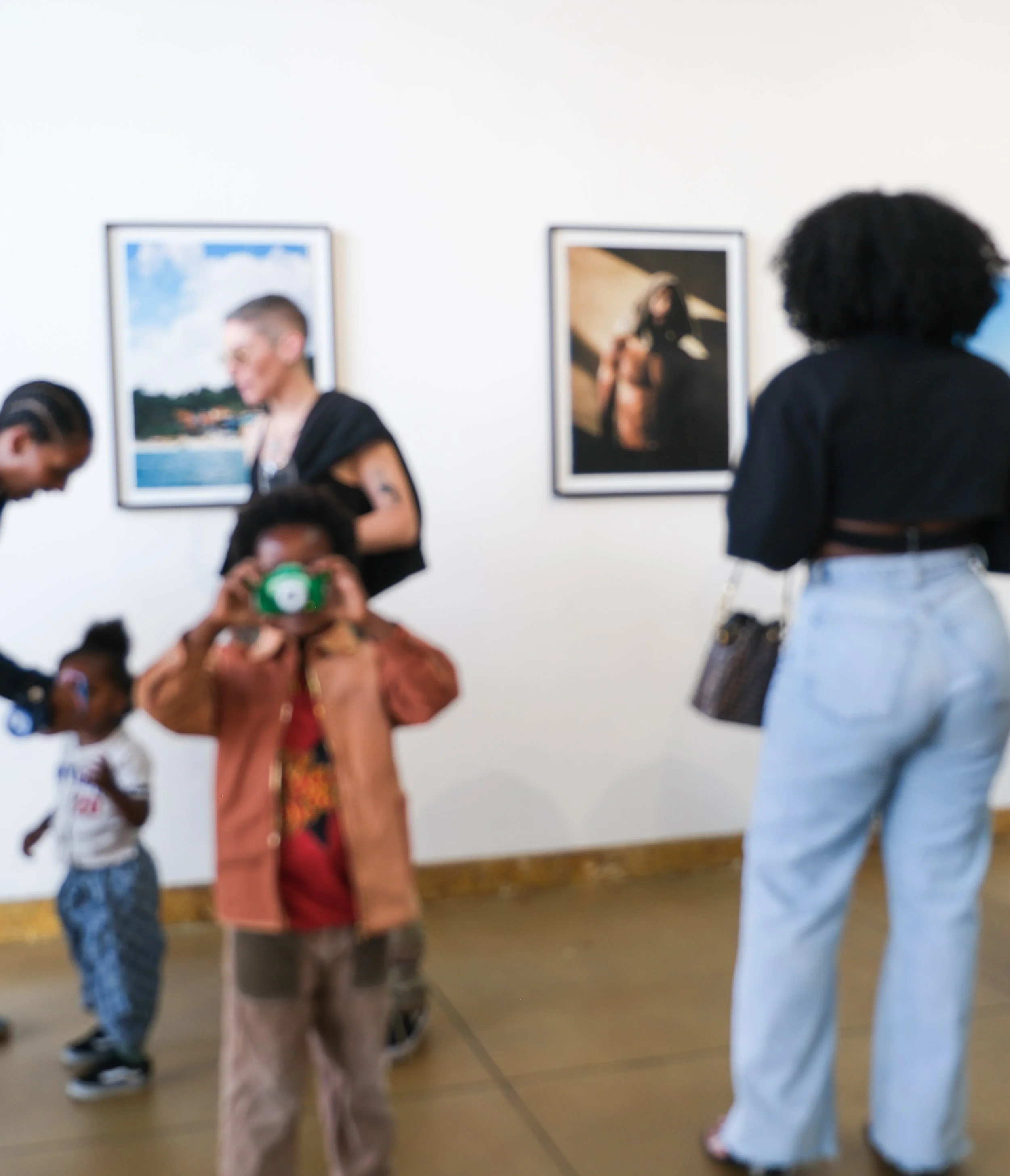 People gathered in an art gallery, viewing framed photographs on the white wall. The focus is on a person wearing a black top and having curly hair, with several others in the background.