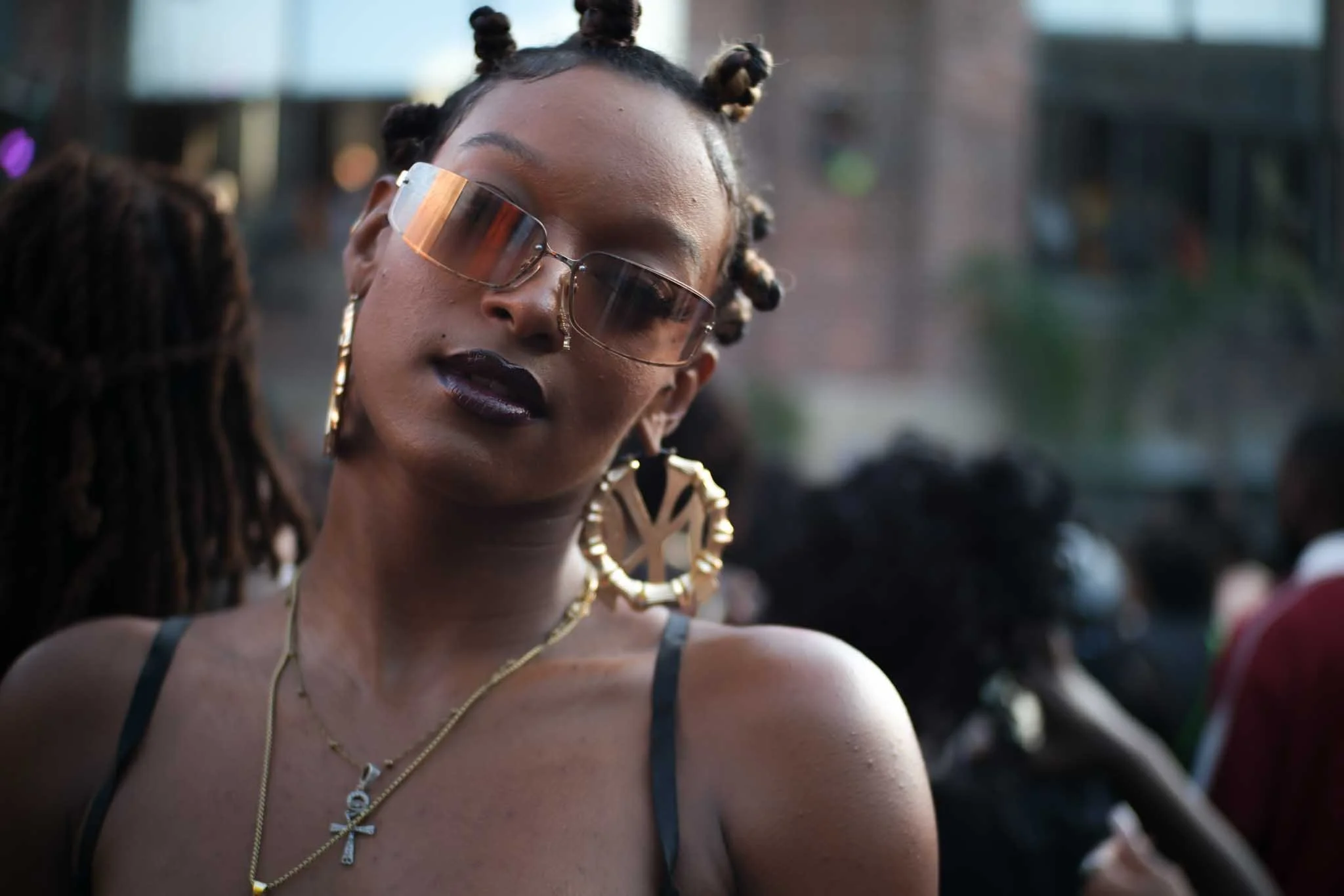 A woman with dark lipstick, wearing large sunglasses, gold hoop earrings, and layered gold necklaces, with her hair styled in bantu knots, outdoors in an urban setting.