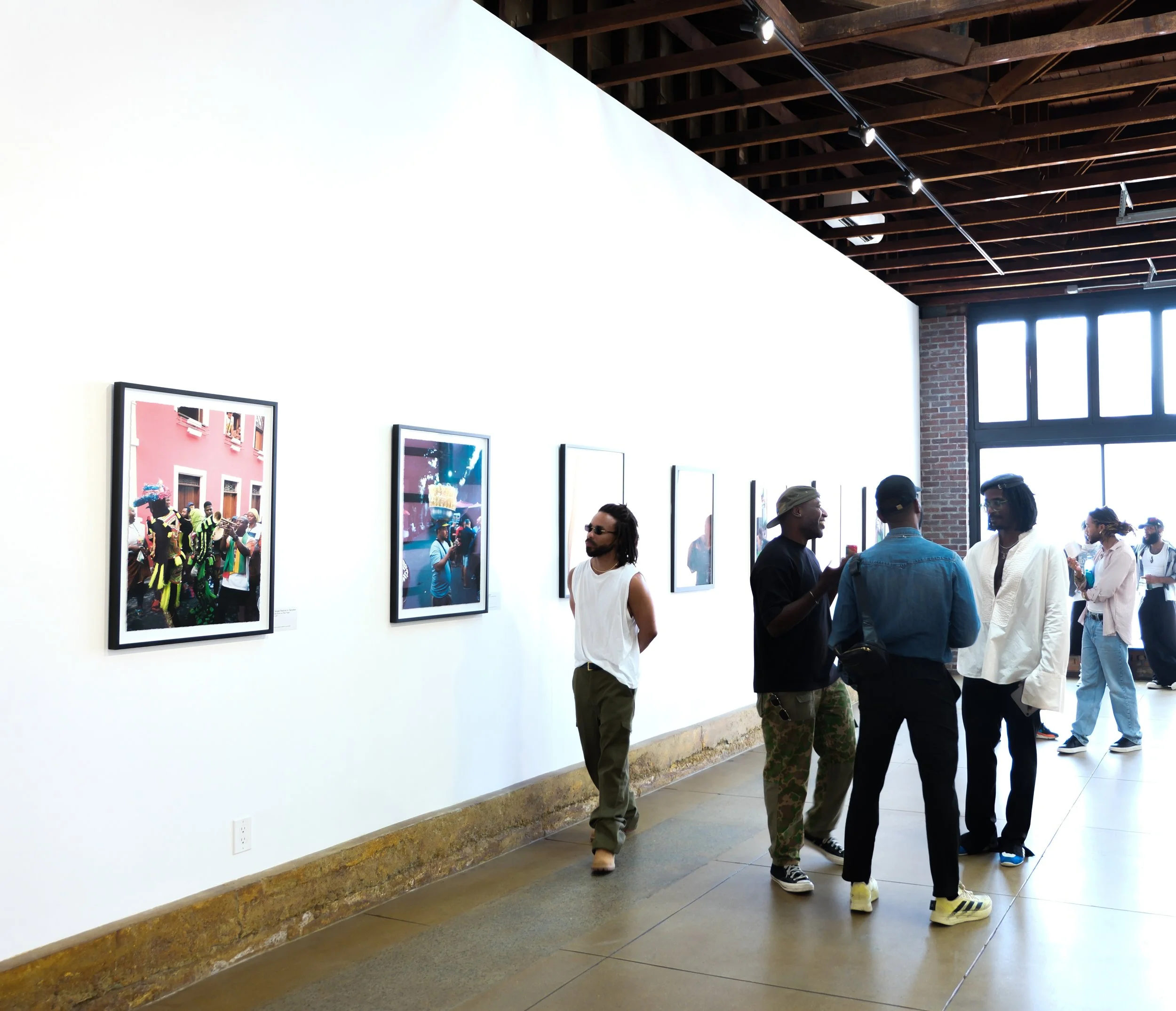 People walking and conversing in an art gallery with framed photographs hanging on a white wall and a brick wall near a window with natural light.