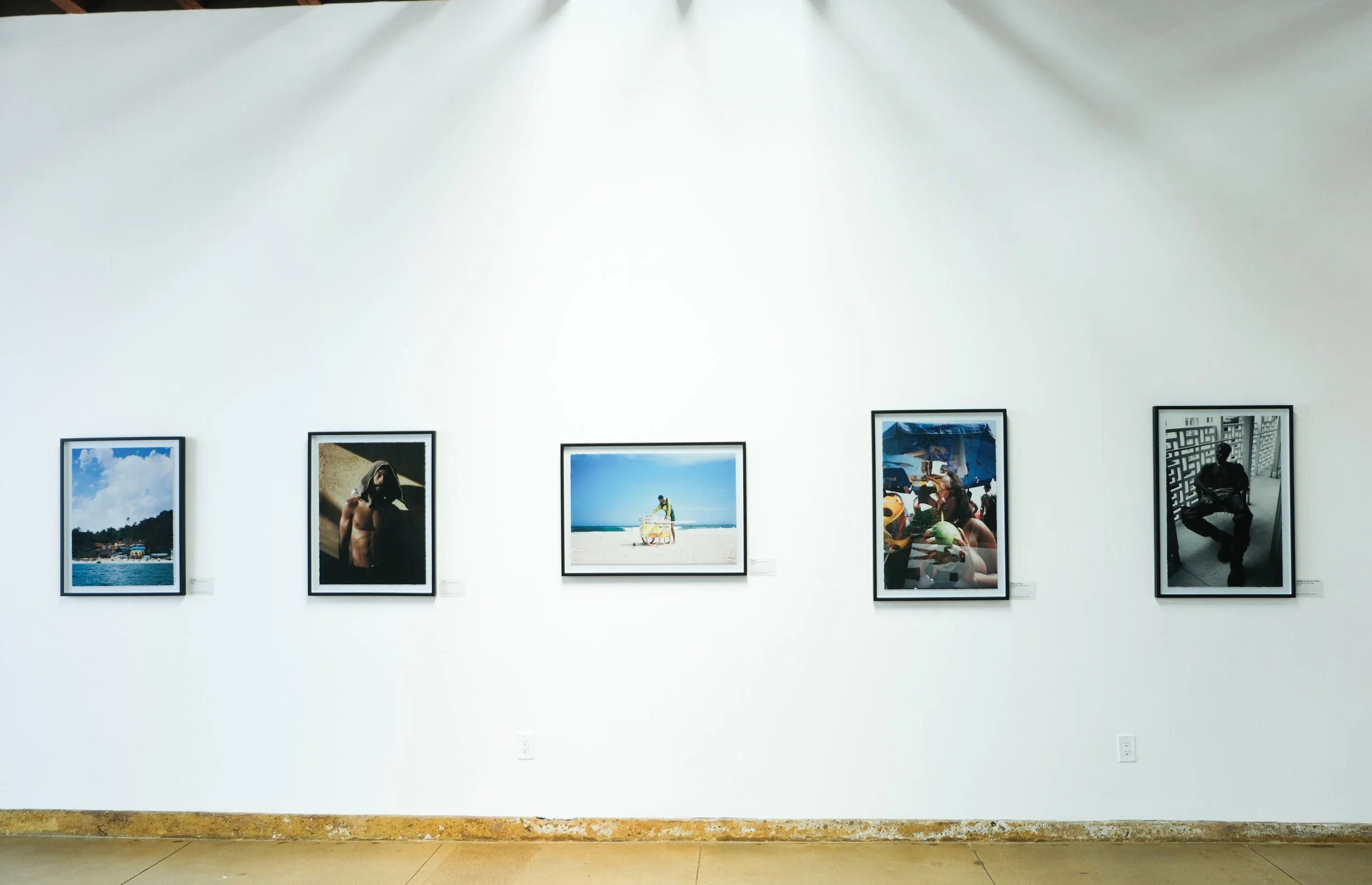 Gallery wall with six framed photographs of people and scenic landscapes displayed on a white wall in an art gallery.