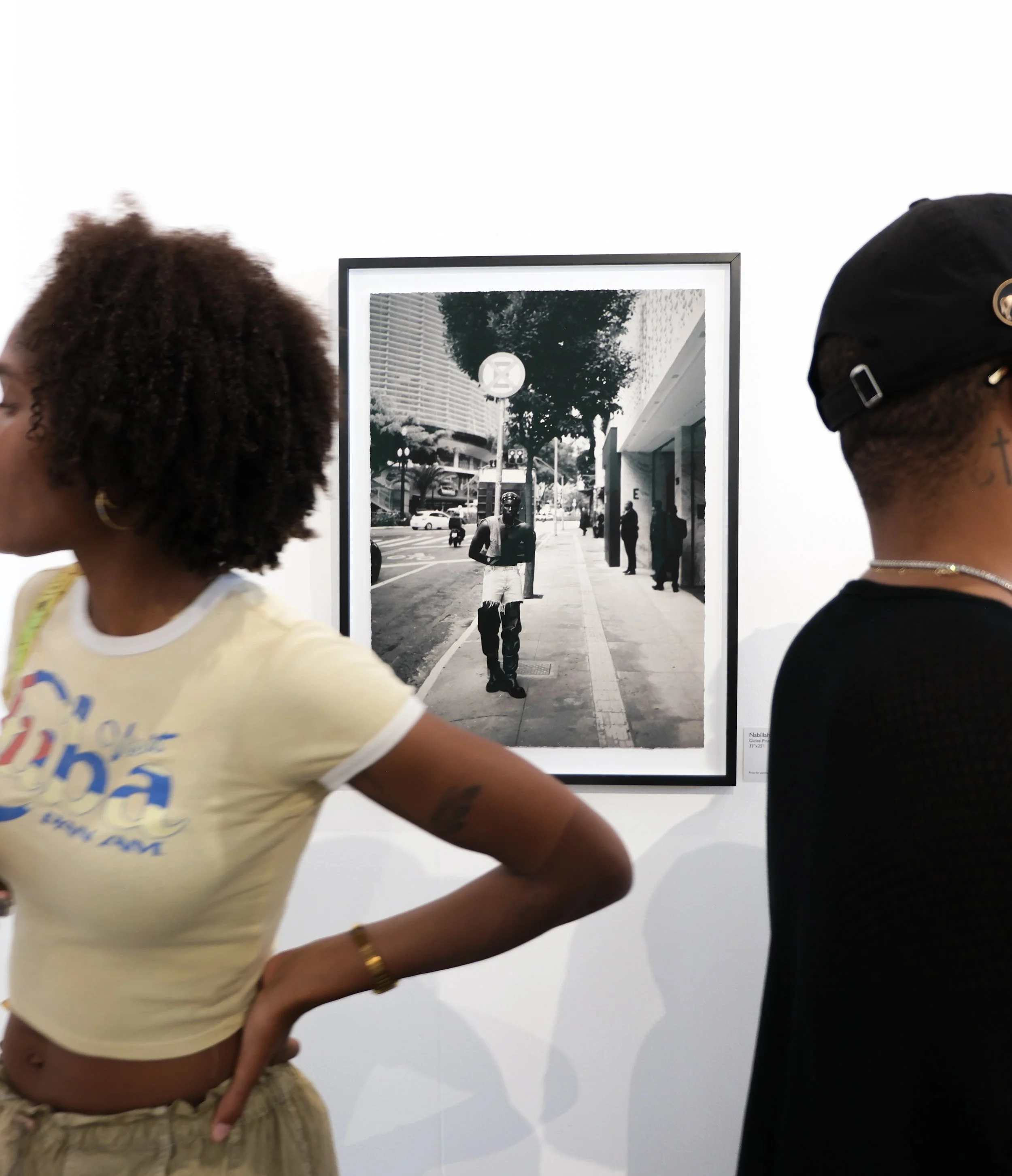 Two women observing a black-and-white photograph in an art gallery. The person on the left has curly short hair and wears a yellow shirt. The person on the right has short hair, is wearing a black cap, glasses, and a black shirt. The photo they are v