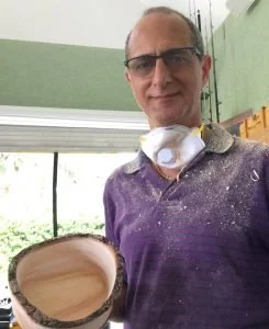 Man holding a piece of wood, wearing glasses and a purple shirt with a dust mask around his neck, inside a workshop or garage.