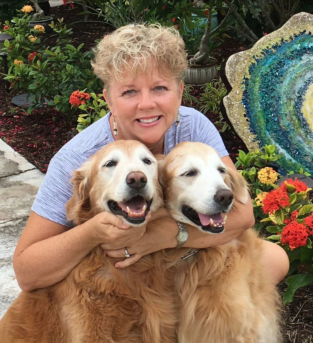 A woman with short, curly blonde hair smiling while hugging two happy golden retriever dogs in a garden with colorful flowers and a decorative stone art piece.