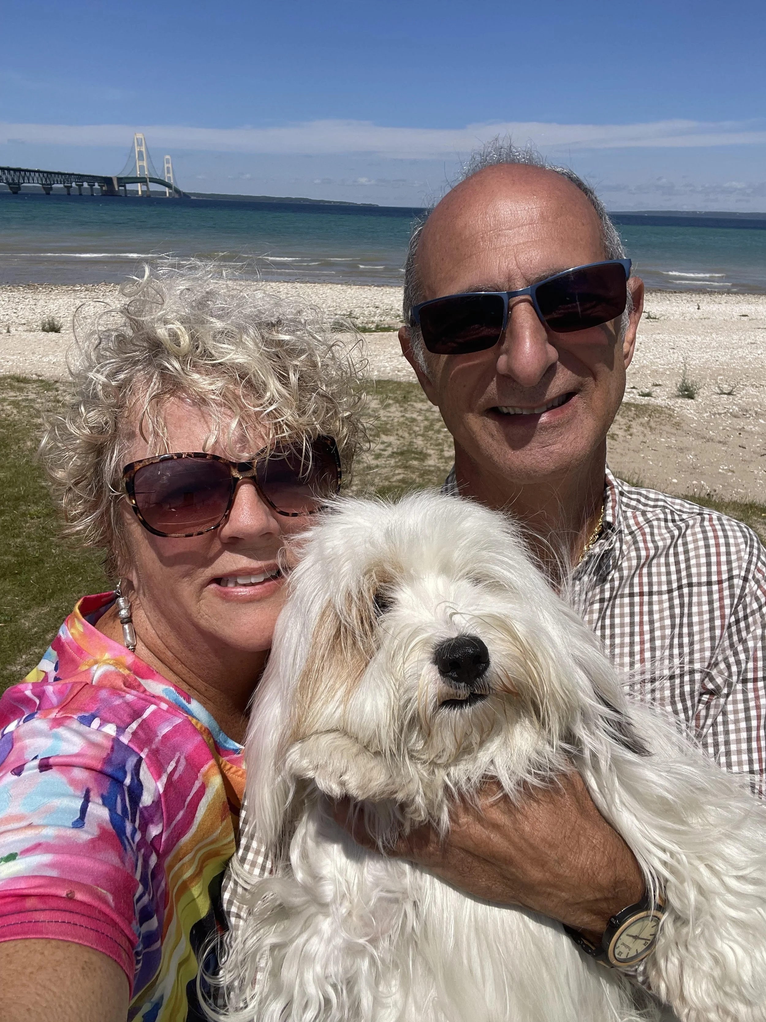 A smiling elderly man and woman with dark sunglasses taking a selfie on a beach, holding a fluffy white dog in front of them. The background features blue sky, water, and a bridge.