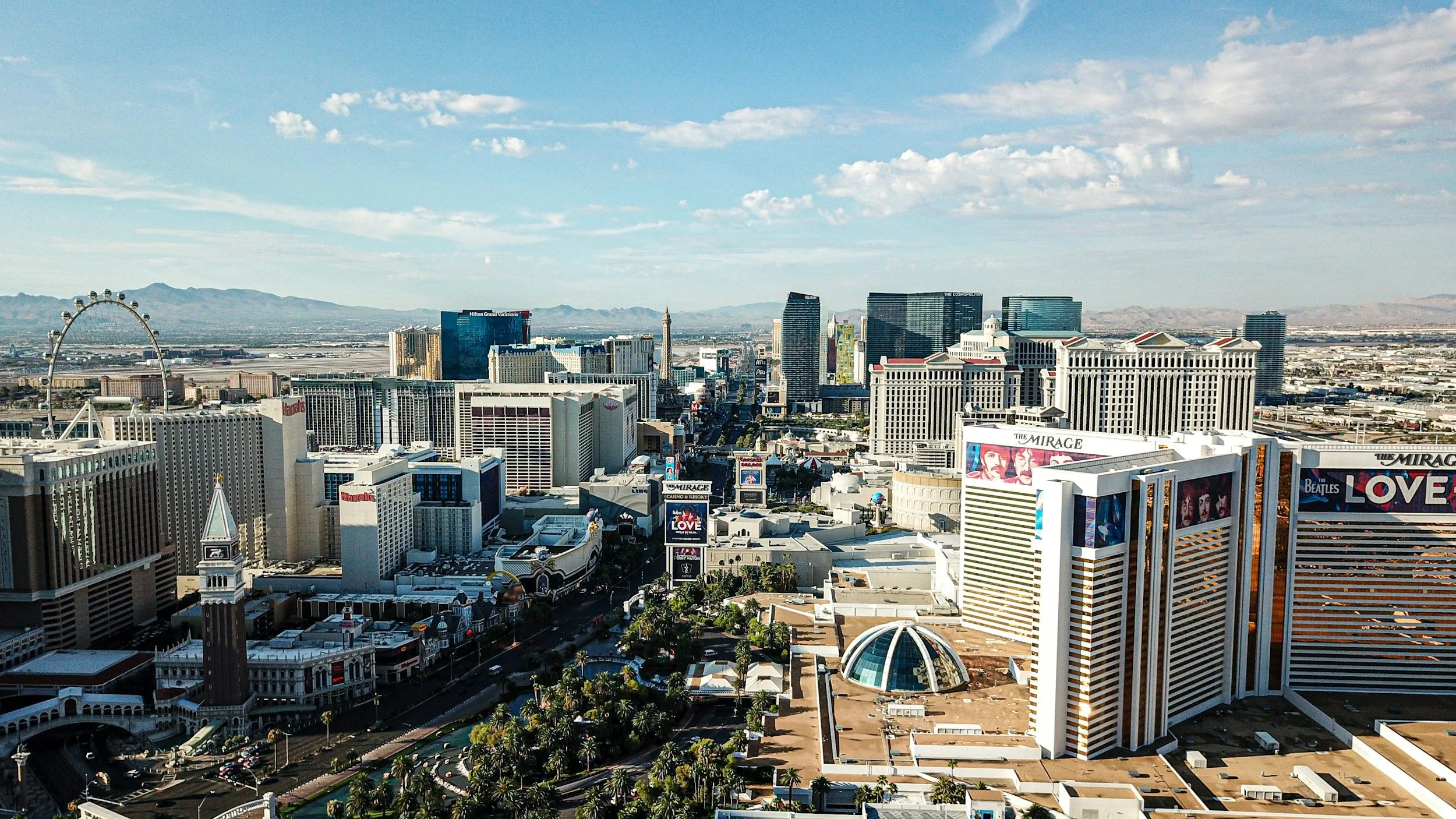 A panoramic view of the Las Vegas Strip with numerous high-rise hotels and casinos, including the High Roller observation wheel to the left and the New York-New York Hotel & Casino in the foreground, under a partly cloudy sky.