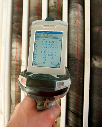 Hand holding a portable water testing device displaying water quality measurements, with several black pipes sealed with red tape in the background.
