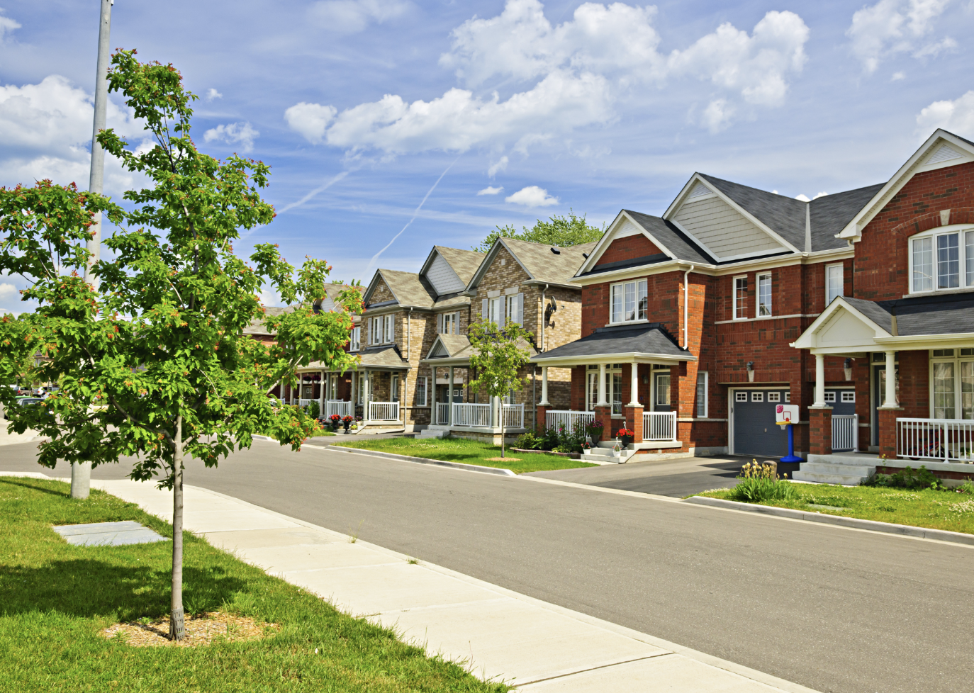 A row of modern, closely spaced suburban houses with front porches and well-maintained lawns. There is a green tree in the foreground, a paved street, and a partly cloudy sky.