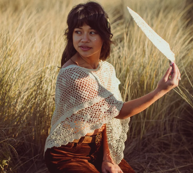Woman sitting in a field of tall grass, holding a white feathered fan, wearing a crochet top and brown pants, with shoulder-length dark hair and a small hair clip.