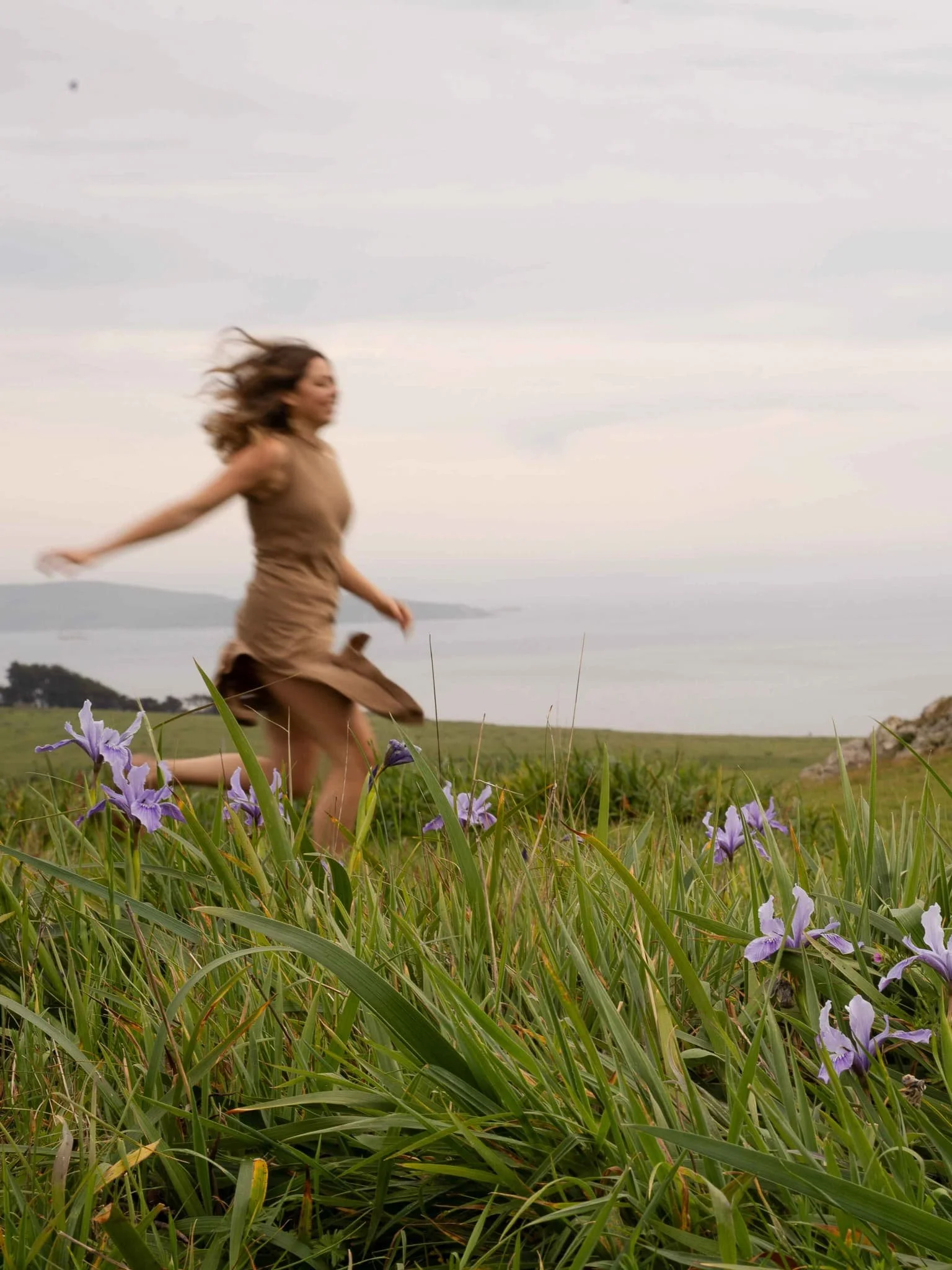 A woman in a tan dress running through a lush green field with purple flowers, with rolling hills and cloudy sky in the background.