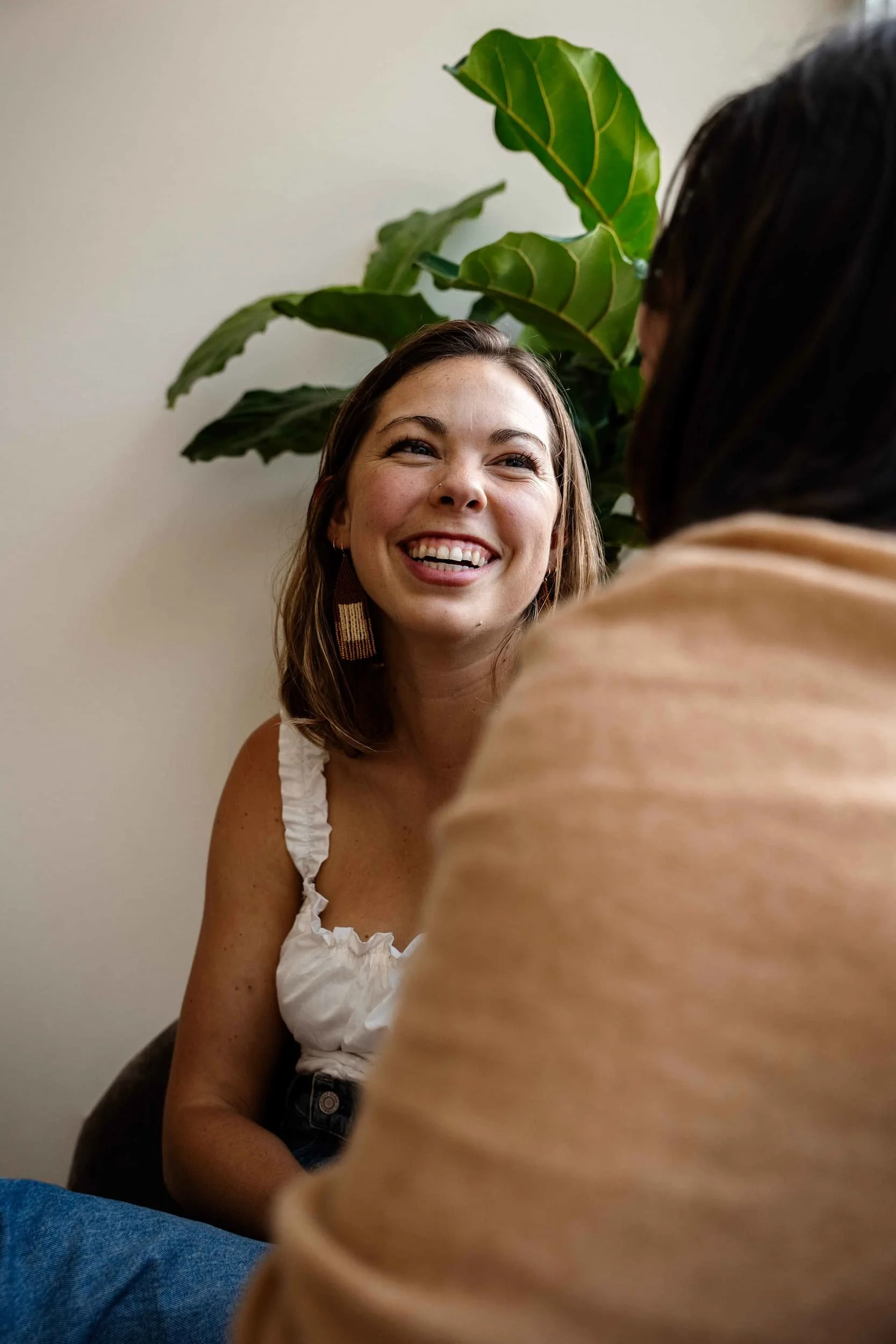 A woman with shoulder-length brown hair and earrings is smiling and looking at another person, partially visible, in front of her. They are indoors with a large green leafy plant behind her, and the woman is wearing a white ruffled top.