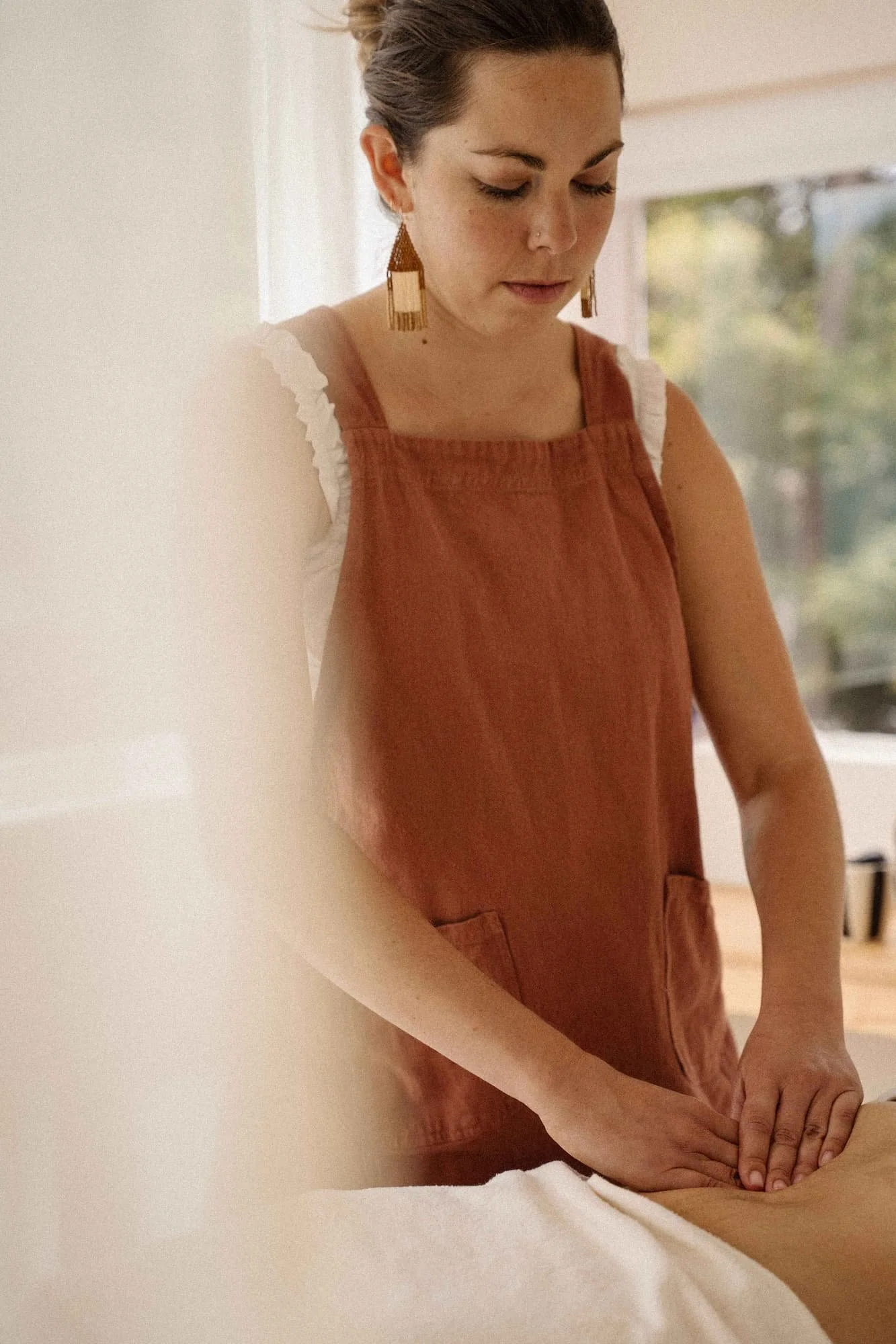 A woman with brown hair in a bun, wearing large earrings and an orange apron, appears to be giving a massage or similar treatment to a person lying on a table, in a softly-lit room with a window in the background.