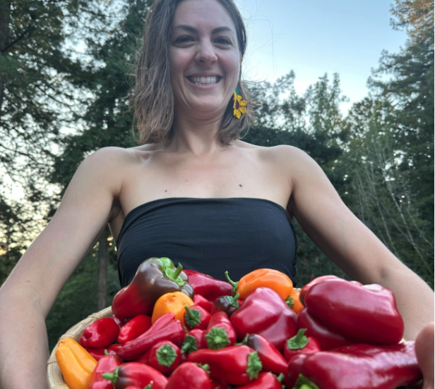 Smiling woman holding a basket of red and orange peppers outdoors, with trees and a blue sky in the background.