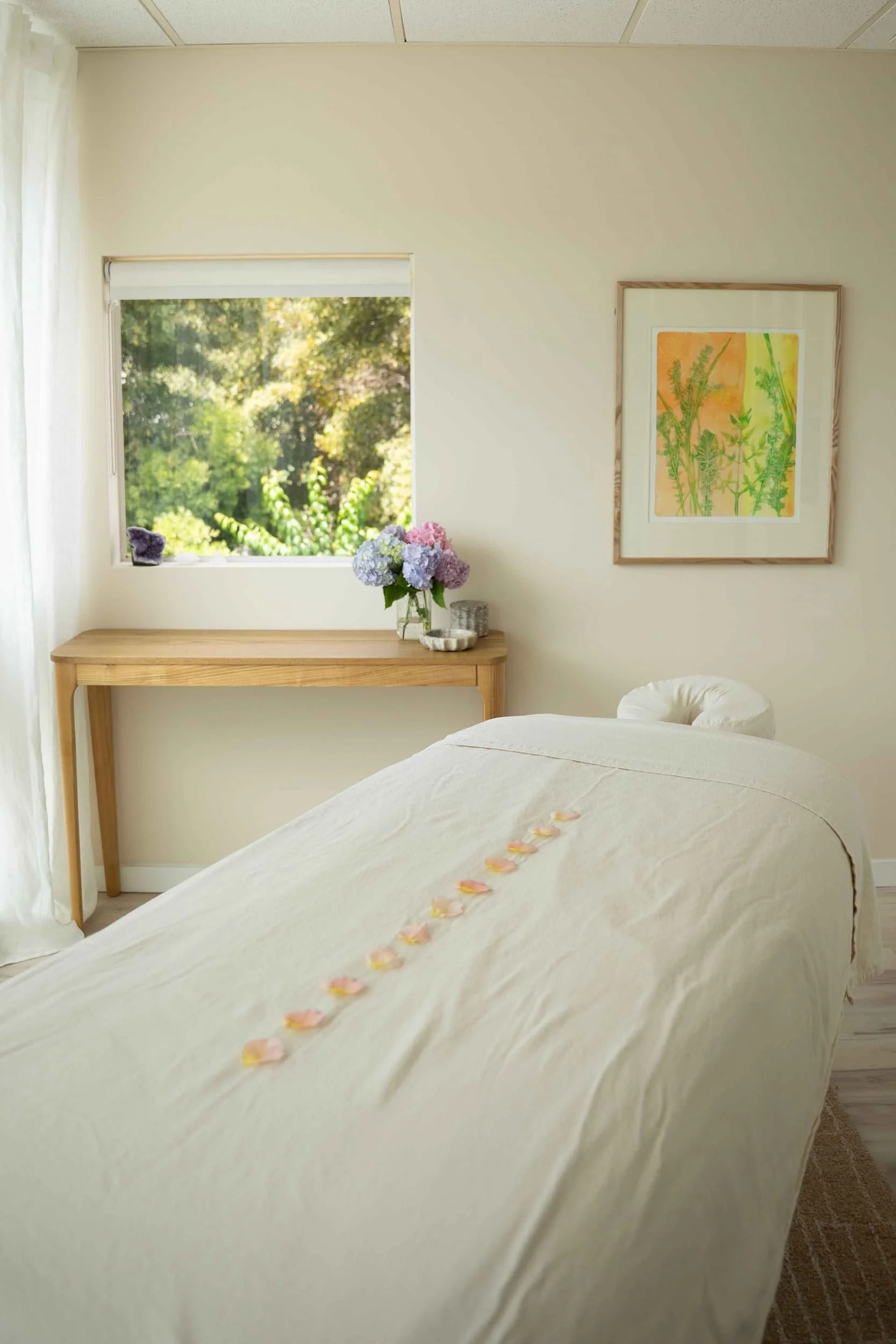 A bedroom with a bed covered in a white sheet and scattered pink and yellow flower petals. A row of petals leads from the bed to the edge. There is a window showing green trees outside, with a wooden table below it holding a vase of hydrangeas and some glasses. On the wall, there is a framed watercolor painting of green foliage and an orange background.