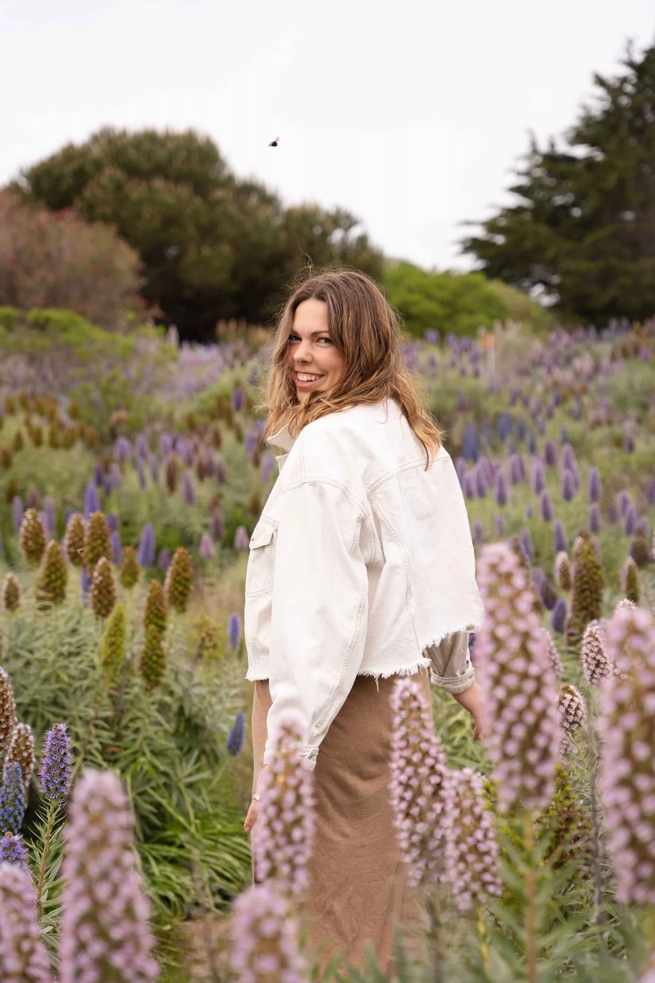Woman with brown hair smiling in a field of purple and pink flowers, wearing a white jacket and a brown skirt, with trees in the background.
