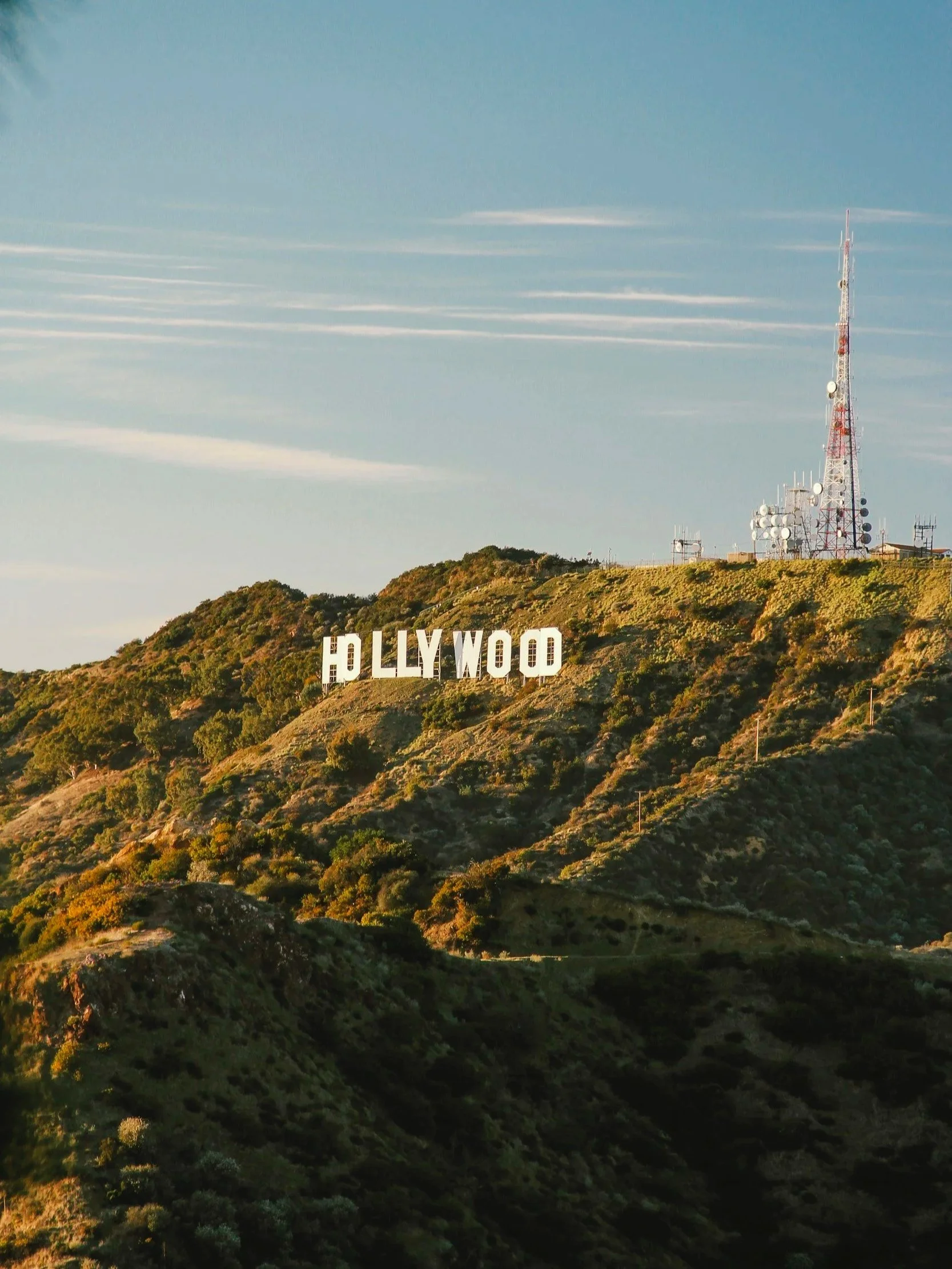 A hill with dense green vegetation and trees, with a radio tower and antennas on top. The Hollywood sign is visible on the hillside, with large white letters spelling 'HOLLYWOOD'. The sky above is mostly clear with a few wispy clouds.