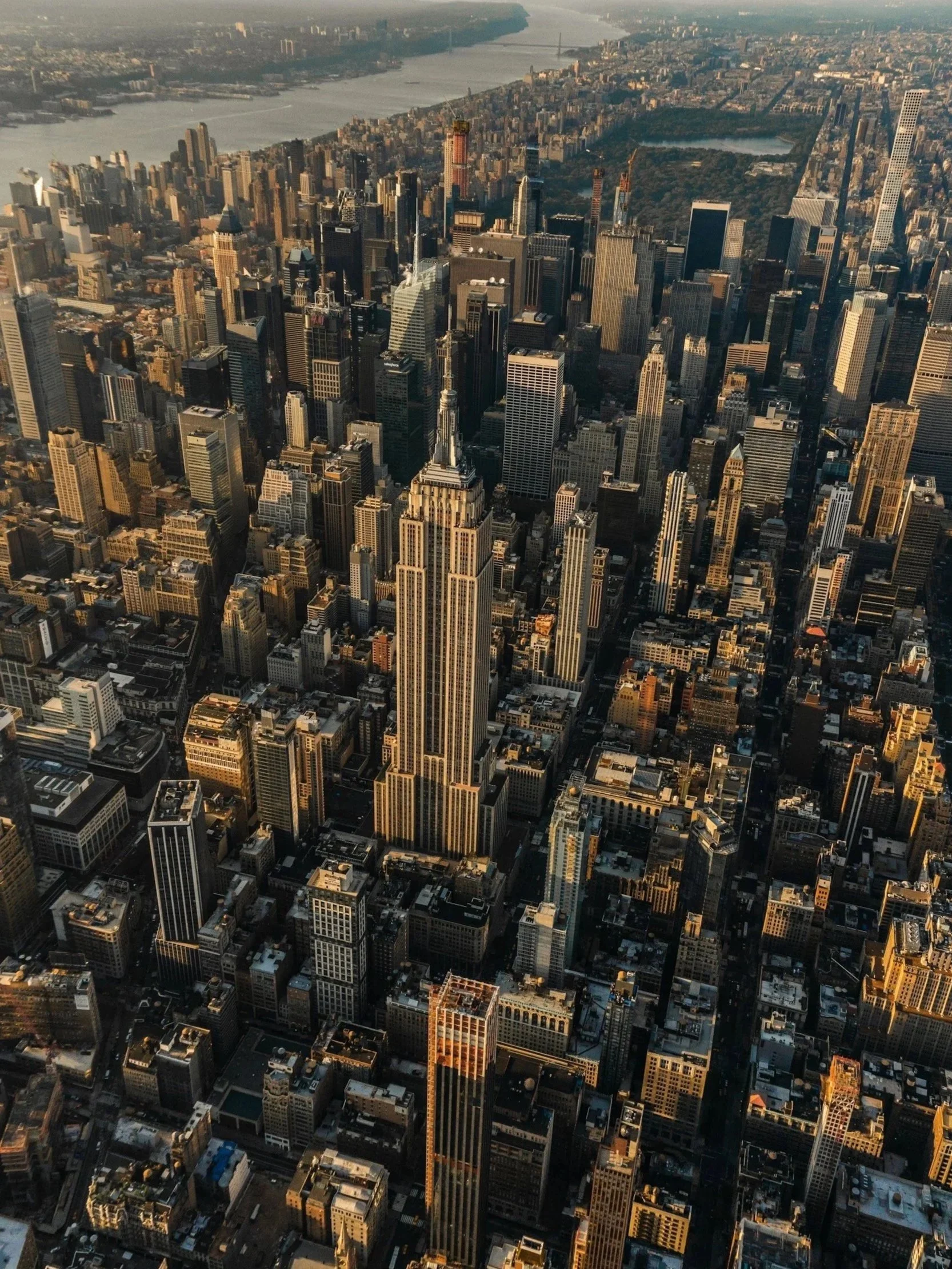Aerial view of the New York skyline with numerous tall skyscrapers, including the Empire State Building, with a river in the background and a bridge across the river.