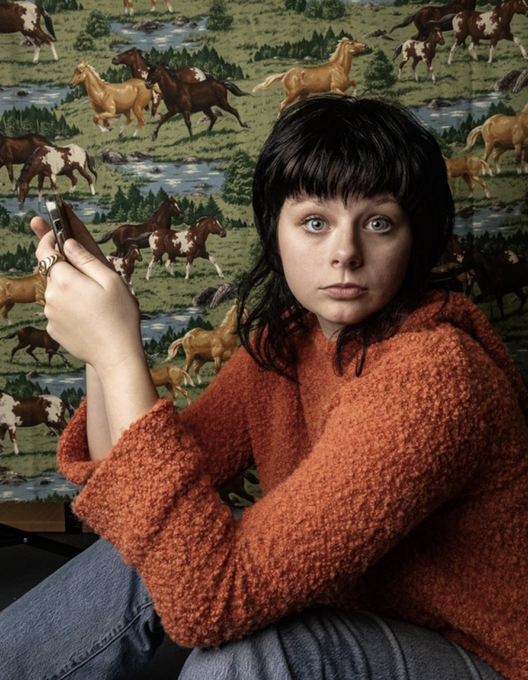 A young woman with black hair and blue eyes, wearing an orange textured sweater, sitting in front of a mural of horses and streams, holding a smartphone.