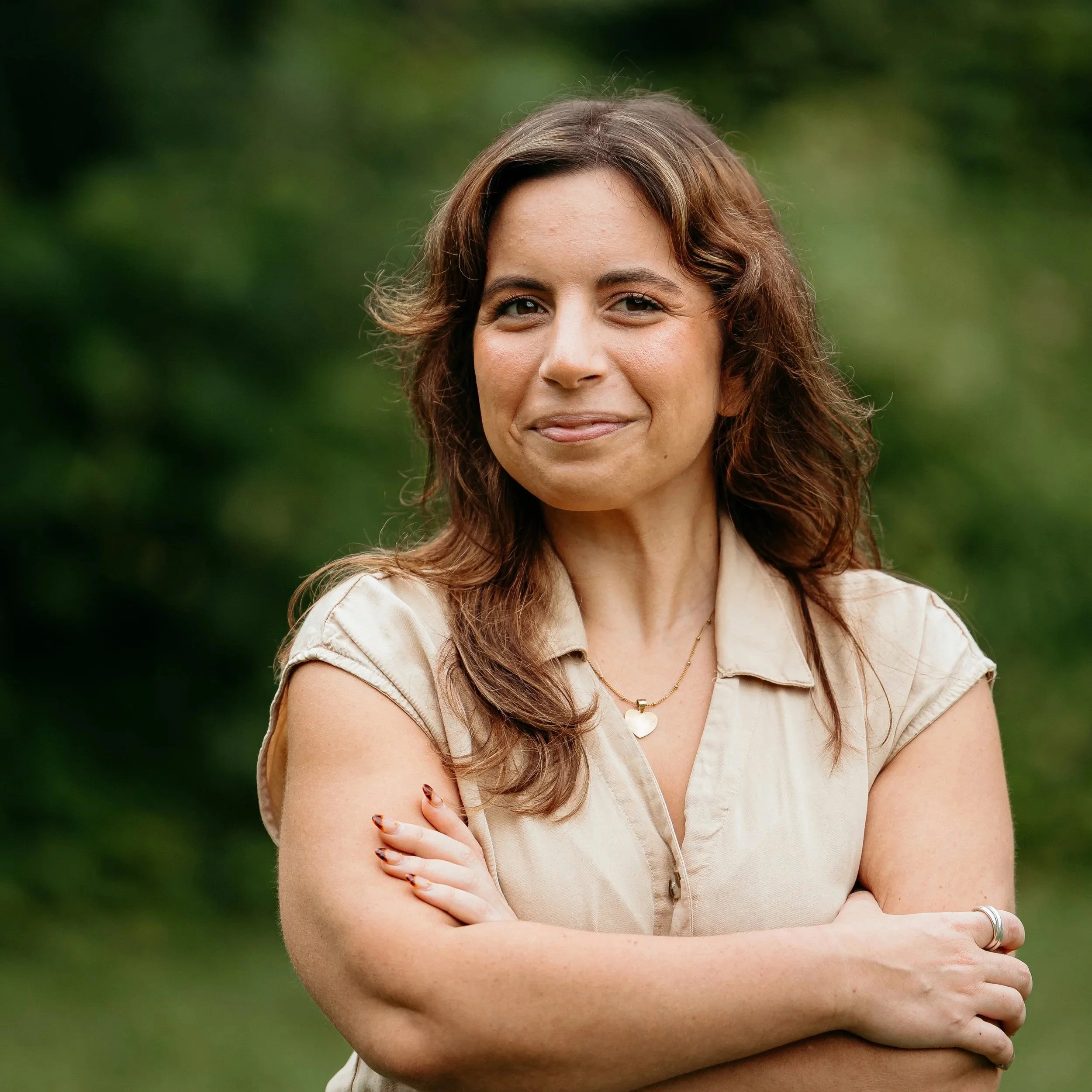 A woman with wavy brown hair and light skin standing outdoors with her arms crossed, wearing a beige shirt with rolled sleeves, a heart-shaped pendant necklace, and a colored bracelet on her right wrist, smiling softly against a blurred green background.