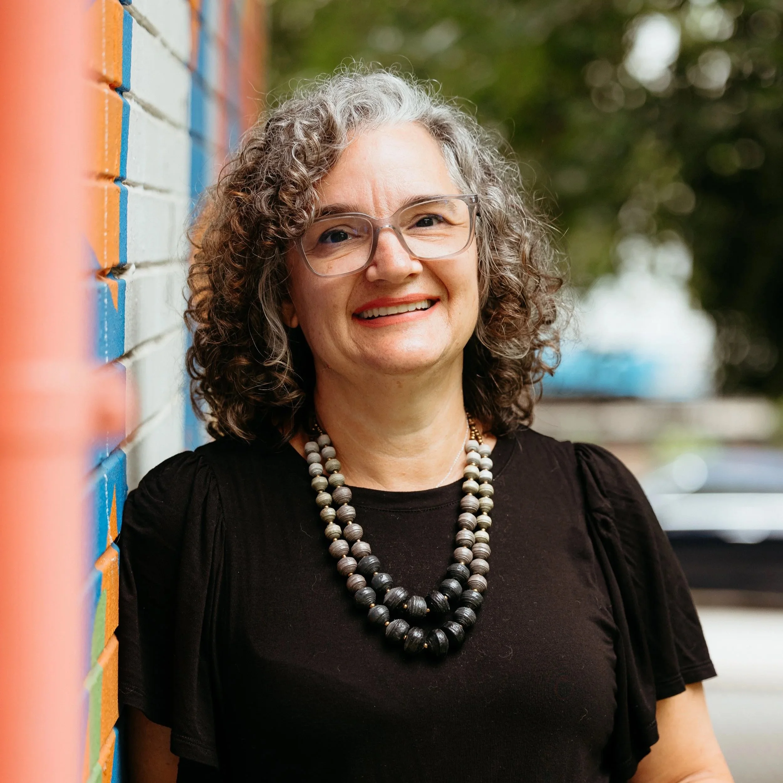 Close-up of a smiling woman with curly gray hair and glasses, wearing a black top and necklace, standing outdoors near a colorful brick wall with blurred greenery in the background.