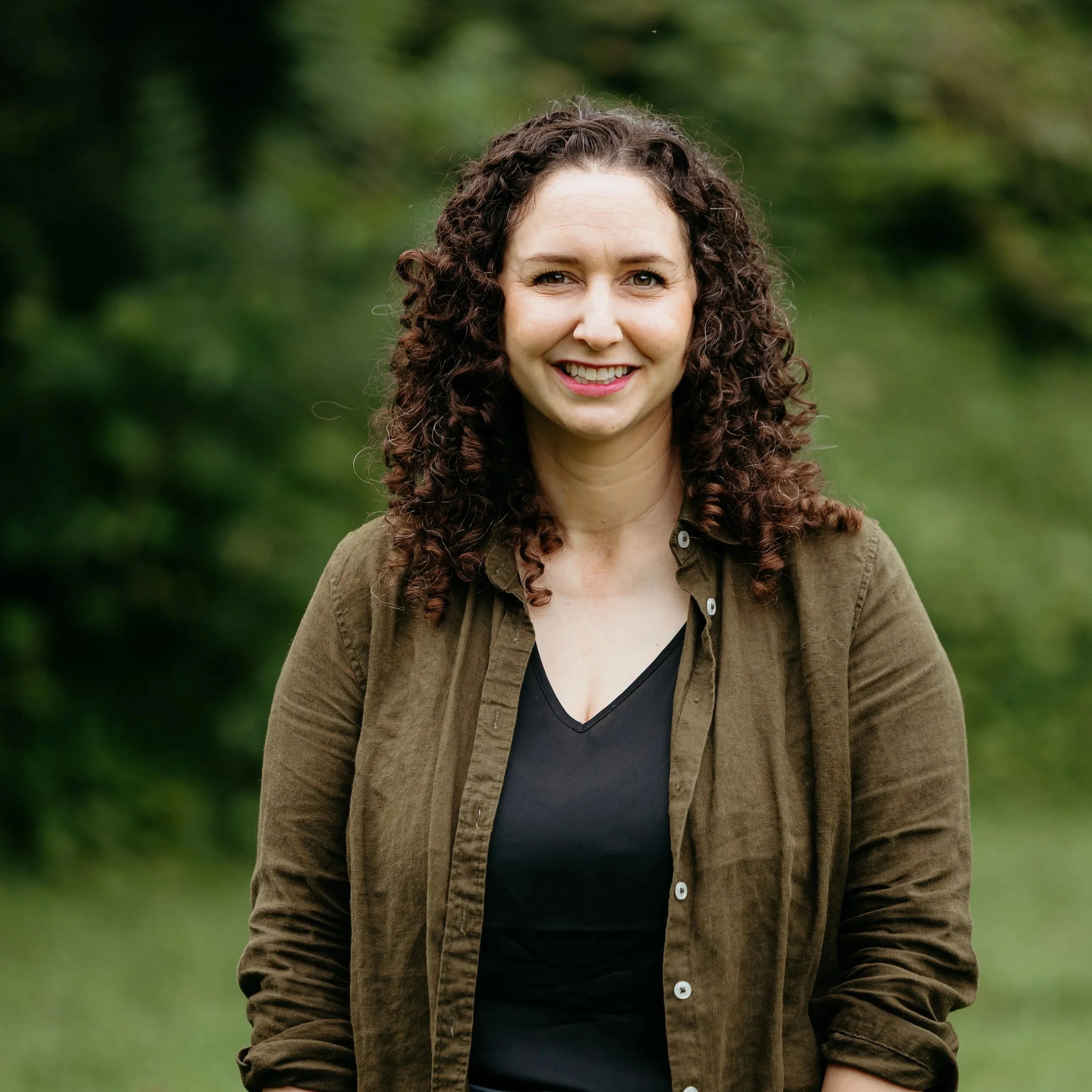 A woman with curly brown hair smiling outdoors, wearing a black top and brown jacket, against a green blurred background.