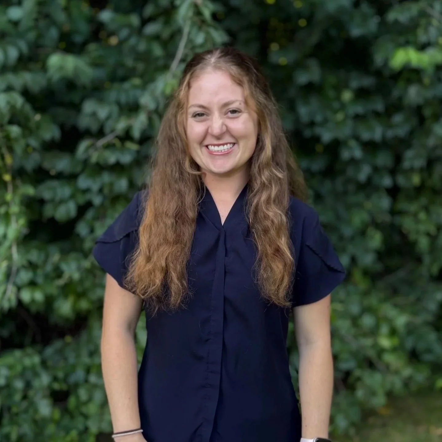 A young woman with long, wavy light brown hair, smiling, wearing a dark blue shirt, standing outdoors in front of green foliage.