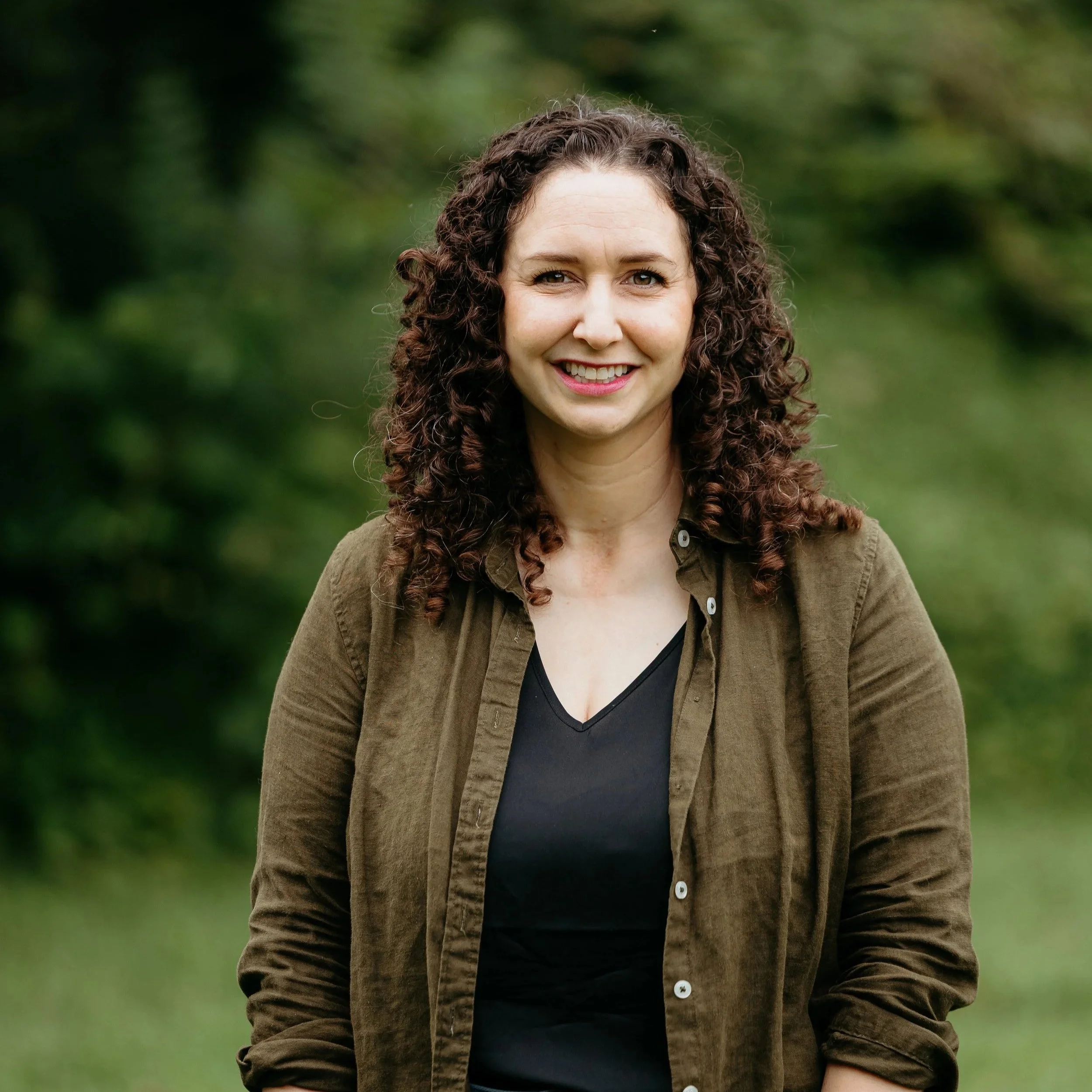A young woman with curly dark hair and a light complexion smiling outdoors with a blurry green background, wearing a brown jacket over a black top.