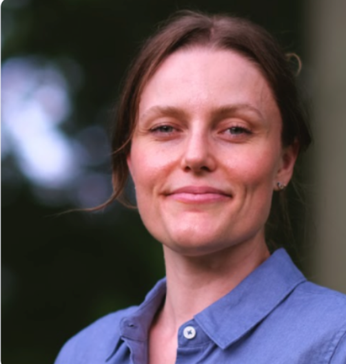 A woman with brown hair wearing a blue collared shirt outdoors with blurred greenery in the background.