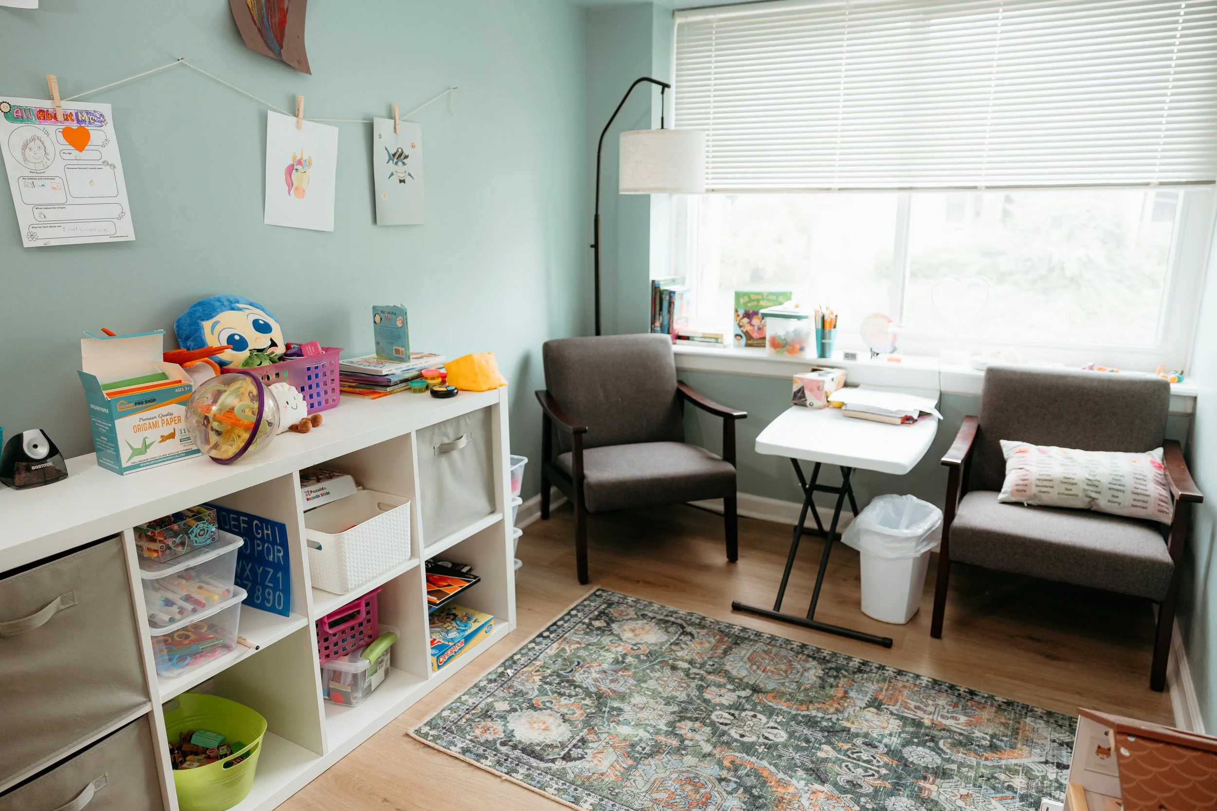 A cozy office or craft room with a bookshelf on the left filled with toys, books, and storage bins. Two grey armchairs and a small white table are near a large window with blinds. The room features a colorful area rug and various decorations on the wall, including drawings clipped to a string.