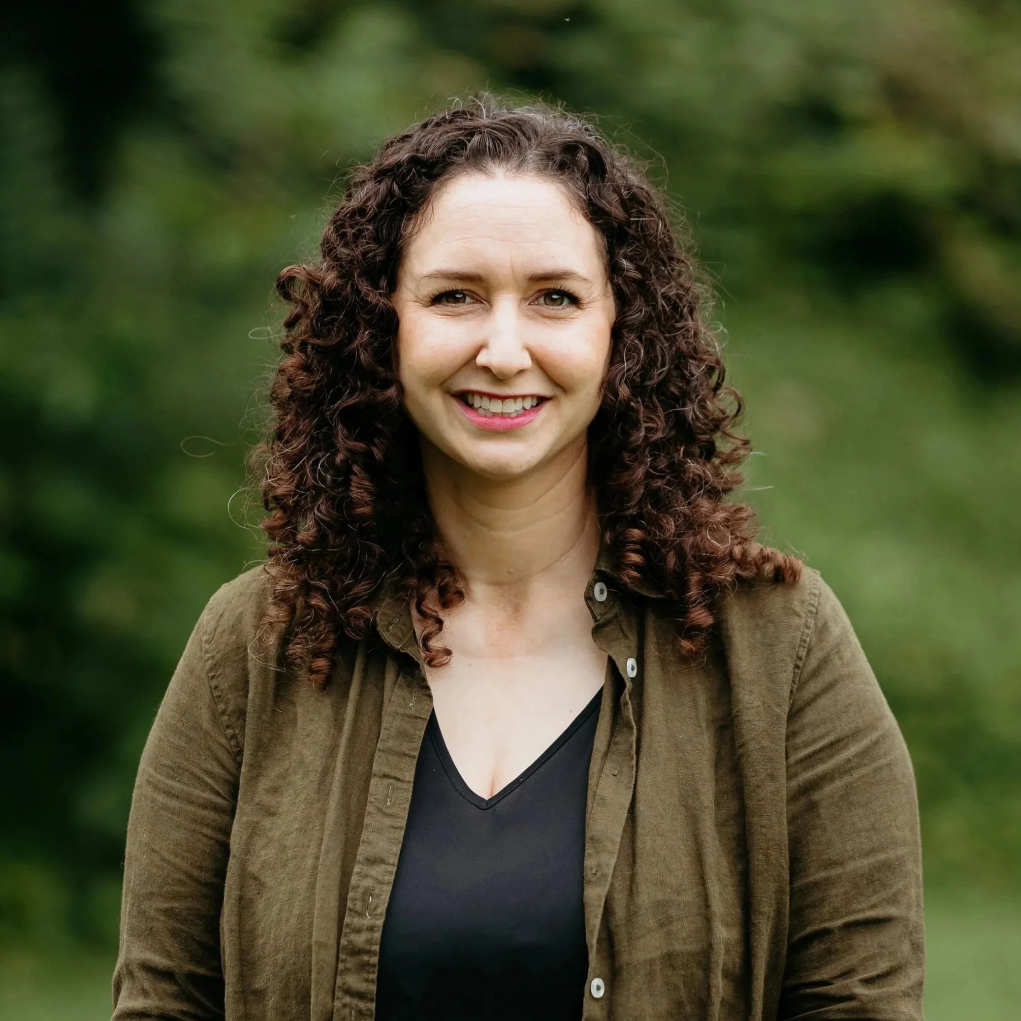 A woman with curly brown hair smiling outdoors with green blurred background, wearing a brown jacket over a black top.