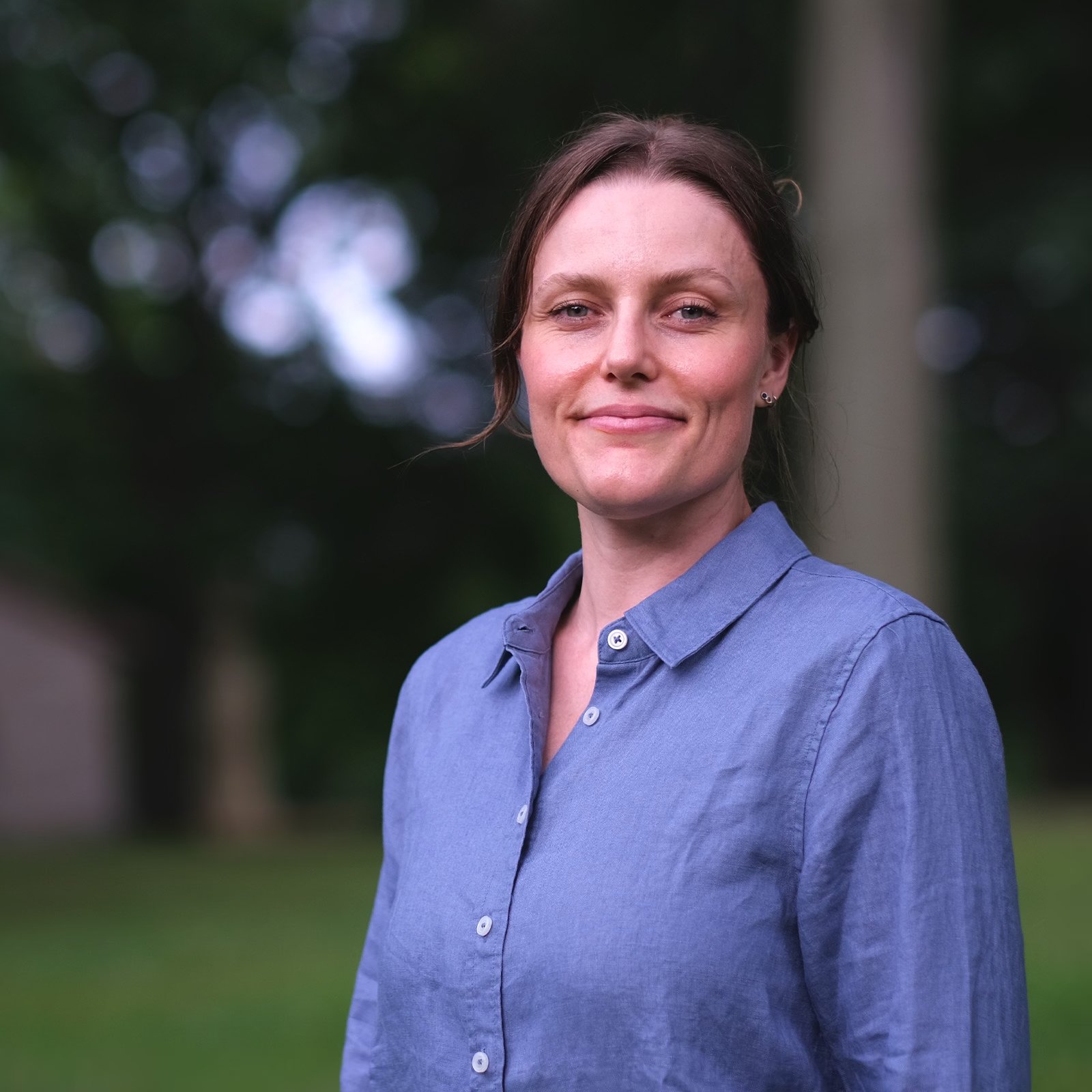A woman with shoulder-length dark hair, wearing a blue button-up shirt, is smiling outdoors with a blurred background of trees and greenery.