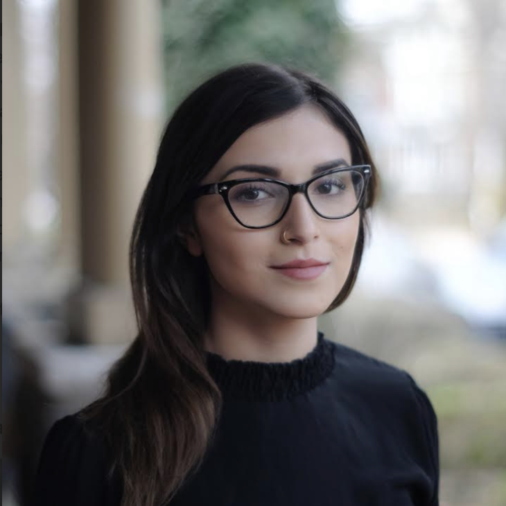 A young woman with long dark hair, wearing glasses and a black top, standing outdoors with blurred background of trees and possibly a building.
