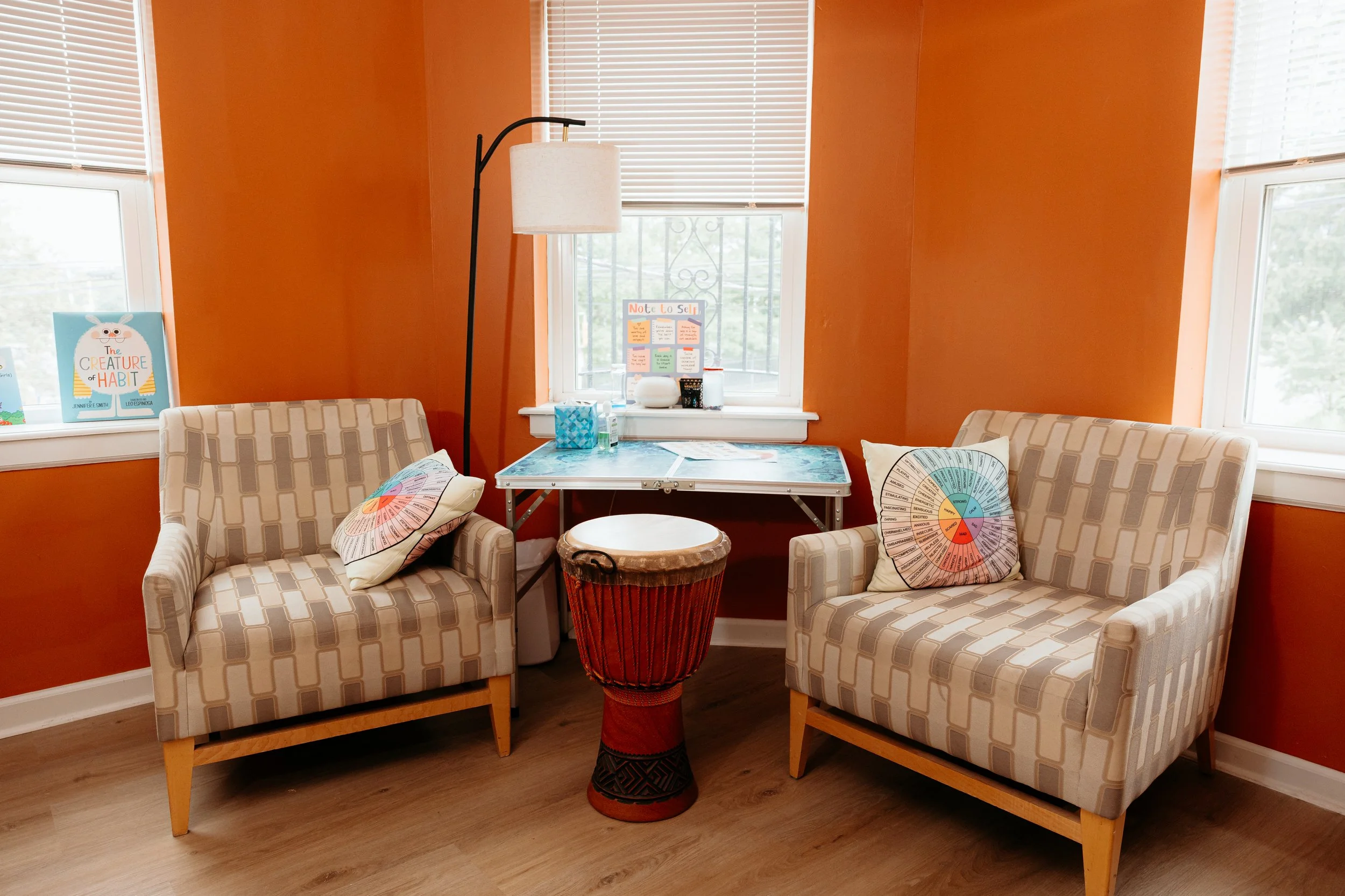 A cozy sitting area with two beige armchairs with geometric patterns, a small round table with a carved wooden base, and colorful pillows. Behind the chairs are large windows with white blinds, and orange walls with a few posters and decorations. A tall floor lamp with a white lampshade is positioned between the windows.